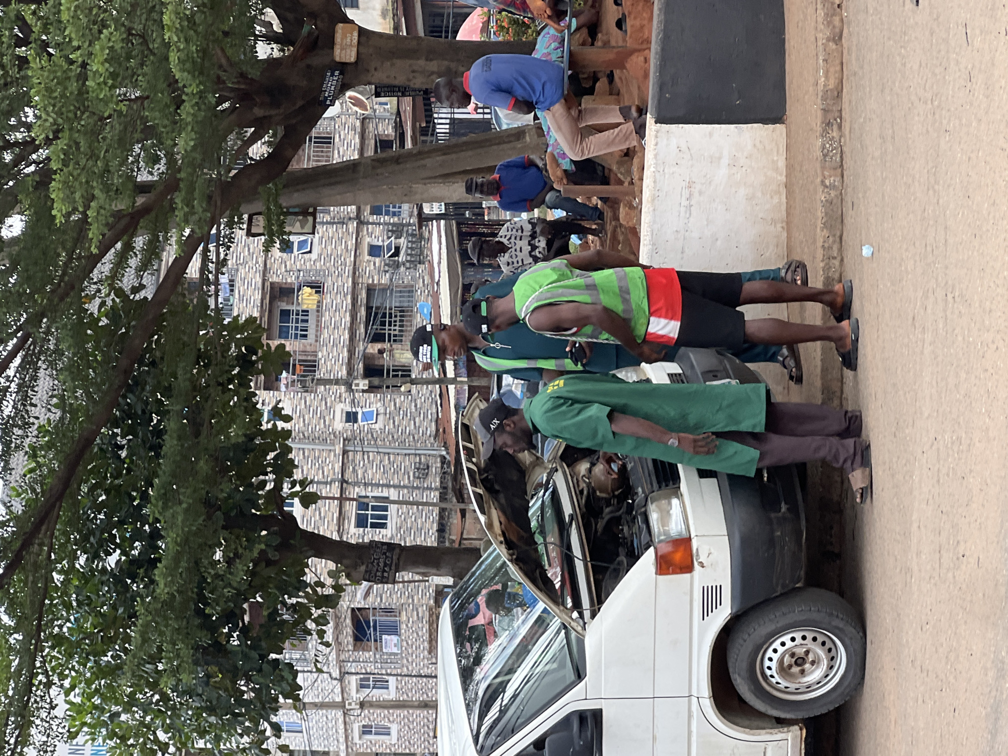 The image shows a group of people gathered around the open hood of a white van, suggesting a car repair. The scene is outdoors, with buildings in the background. The people are dressed casually, with some wearing work vests over their clothing. The overall impression is one of a busy street scene in Nkpor, Nigeria.
The subjects are primarily men, with at least three clearly visible. One man is wearing a bright green vest over a red and white shirt, and black shorts. Another, in a similar green outfit, is leaning over the engine. A third man, wearing a blue shirt and light-colored trousers, is seated in the background. There are other figures partially visible.
The central object is the white van, its hood propped open, revealing the engine compartment. The setting appears to be a street or roadside, with a curb and paved ground. Buildings with multiple windows and balconies are visible behind the people and the van. There's a large tree with dense foliage in the foreground, partially obscuring the view.
The activity is clearly focused on the van's engine, implying a mechanical issue is being addressed. The time of day is difficult to ascertain precisely, but the lighting suggests it could be daytime, possibly late morning or afternoon. There are no obvious cues about the weather, but the general atmosphere seems to be ordinary, without extreme conditions.
A sign is visible on the tree trunk, but the text is too small and blurry to read clearly. However, a few letters and shapes can be discerned, and one word appears to be "PLUMBER," suggesting a connection to a service business, though it's not directly related to the car repair. Another sign on the same tree has some text written on it, but it's also illegible.