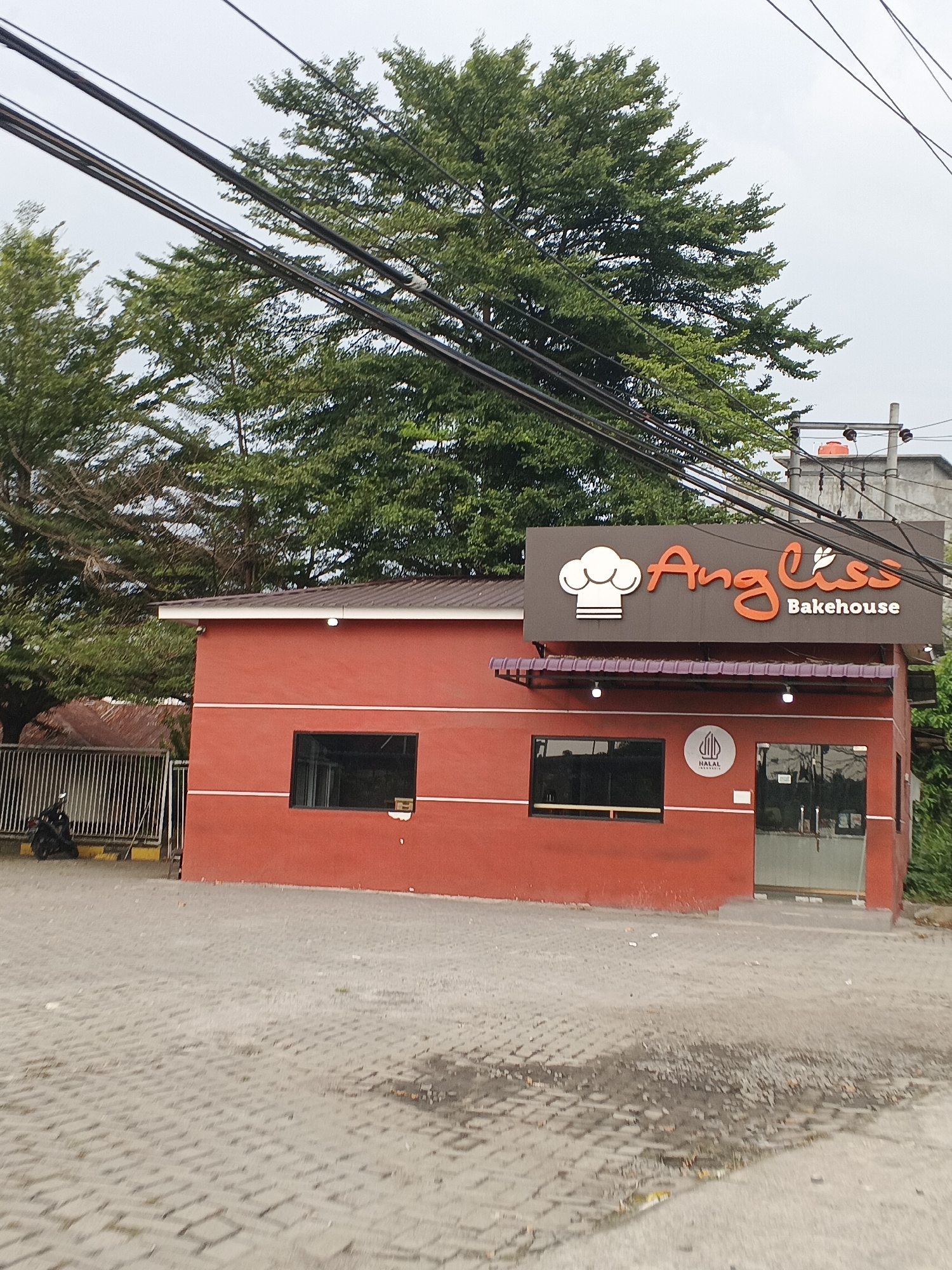 The image captures a bright red building with a dark brown sign that reads "Angliss Bakehouse" in orange lettering and a white chef's hat logo. The building has several dark-framed windows and a glass door. A small circular logo, appearing to be a halal certification, is visible on the facade. The building is situated on a paved area with a patterned brick surface. In the background, lush green trees dominate the upper portion of the frame, with power lines crisscrossing in front of them. A metal fence and a motorcycle are visible to the left of the building. The sky is overcast. The location is identified as Tg Morawa B, Indonesia.