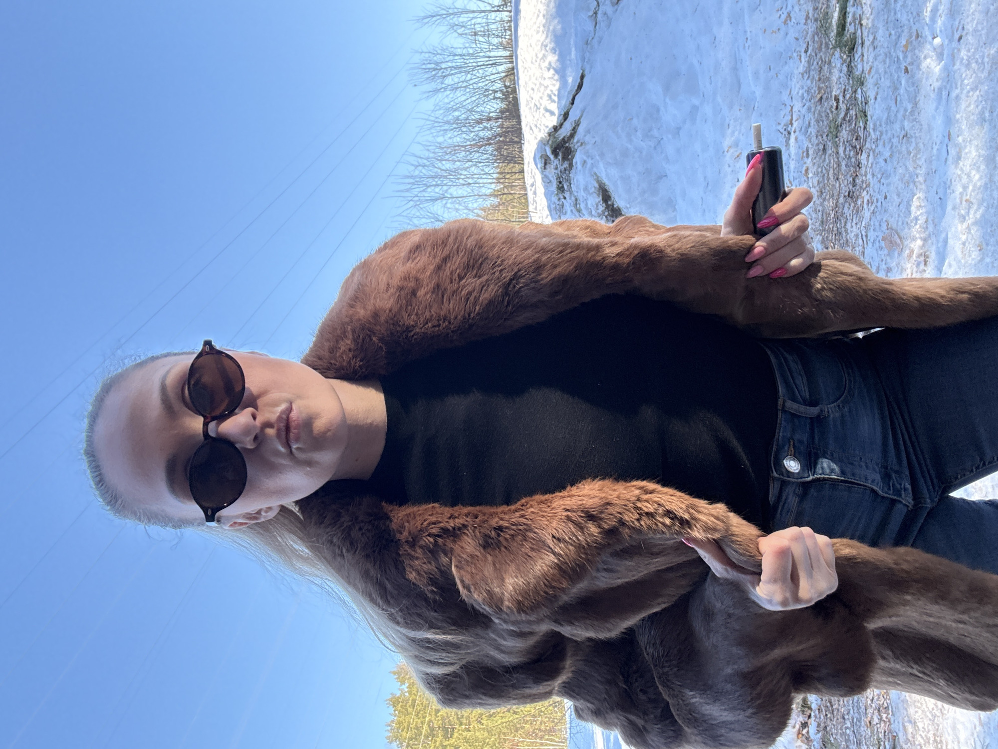 A female individual stands outdoors in Noviy, Russia, under a clear blue sky traversed by utility lines. She is viewed from the waist up, facing the camera with a neutral expression and slightly pursed lips. She wears dark, round-framed sunglasses, a dark brown fur coat over a black long-sleeved garment, and dark blue jeans. Her light-colored hair is visible around her face. Her fingernails are painted bright pink. In her right hand, she holds a black cylindrical object with a light-colored tip, consistent with an electronic vaping device. Her left hand holds the lapel of her fur coat. The background consists of snow-covered ground with exposed earth patches, evergreen trees to the left, and bare deciduous trees on a snow-dusted hill to the right. The scene is brightly lit by natural sunlight.