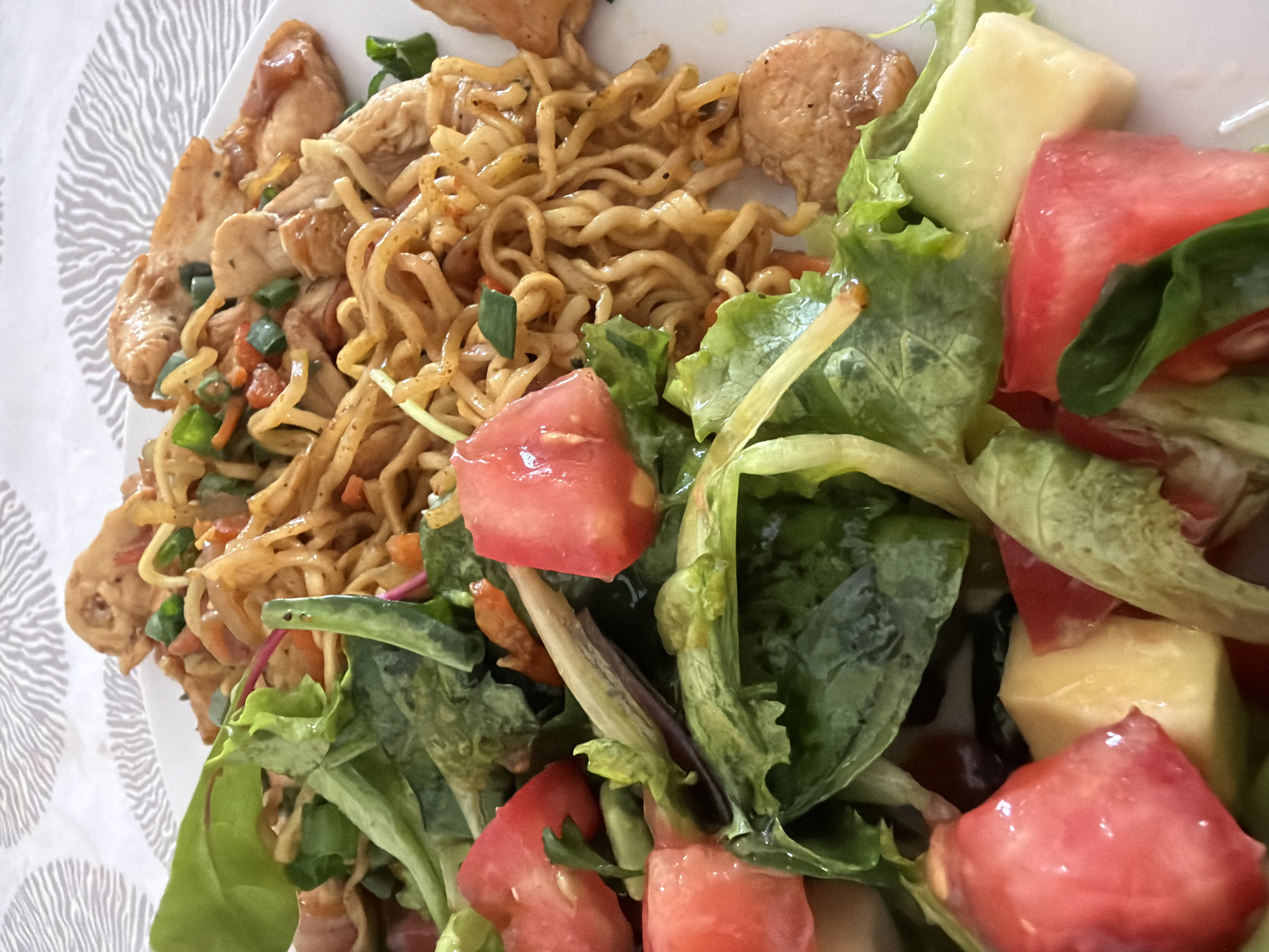 A close-up, eye-level shot shows a white plate of food. The food consists of a portion of stir-fried noodles with chicken and vegetables, and a side salad with tomatoes and lettuce. The noodles are thin and appear to be a savory brown color, mixed with diced carrots, green onions, and pieces of chicken. The salad is a mix of fresh green lettuce leaves, diced red tomatoes, and some lighter colored diced pieces that could be cucumber or cheese. The background is a textured white tablecloth. There are no people or animals visible in the image, and no specific geographic location can be determined from the visual information provided.