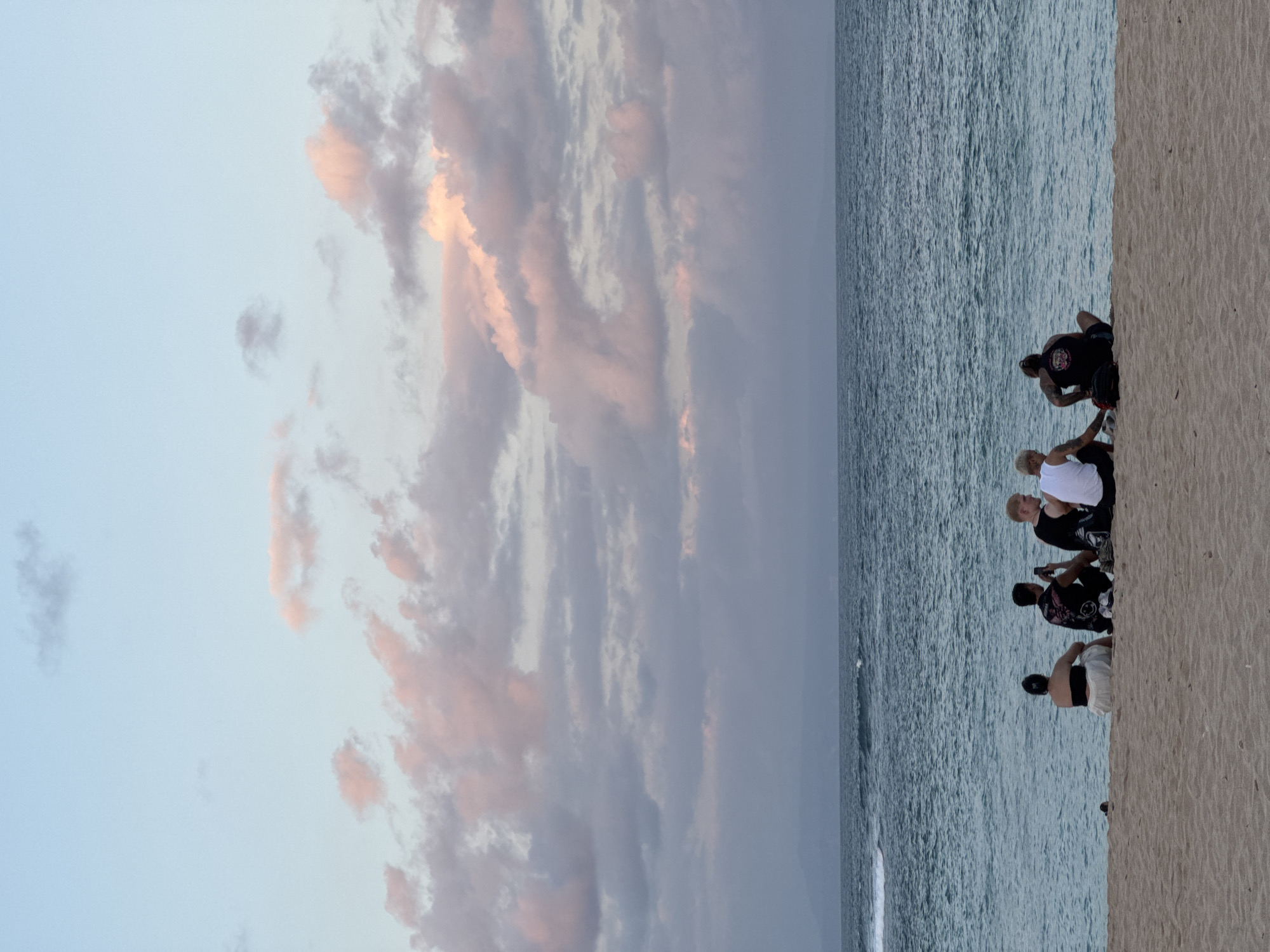 A group of five individuals are sitting on a sandy beach facing the ocean. The sky is a pale blue with scattered clouds tinged with pink and purple, suggesting either sunrise or sunset. The ocean is a deep blue with gentle waves breaking near the shore. The people are dressed casually. From left to right, the first person is wearing a black bandeau top and a white skirt. The second person is wearing a black t-shirt with a graphic print. The third person is wearing a white t-shirt. The fourth person is wearing a black t-shirt with a graphic print. The fifth person is wearing a white t-shirt. The group appears to be engaged in conversation or simply enjoying the view. The location is a beach in Kuta Selatan, Indonesia.