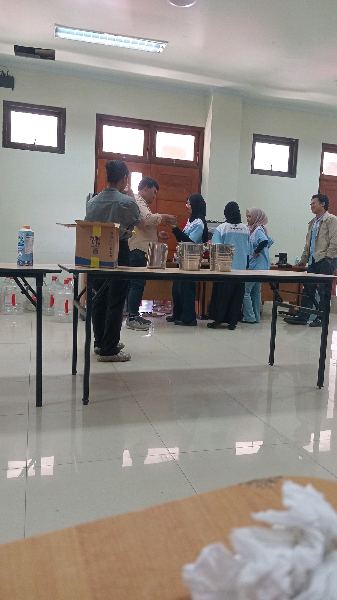 The image depicts an indoor scene, likely a classroom or community hall, with a group of people gathered around a table. The room has light-colored walls, a tiled floor that reflects light, and fluorescent lighting on the ceiling. There are large windows with wooden frames, suggesting a tropical or humid climate.
The main activity appears to be centered around a table where several individuals are interacting with containers and what seem to be beverage ingredients. On the left side of the table, a person in a grey shirt and dark pants is holding a box labeled "Milk Life Barista." Next to it, a carton of "Milk Life" is visible.
Several individuals are wearing blue shirts, some with the logo "SIAP KERJA" visible on the back, indicating a possible training or work-related event. One person in a black hijab and blue shirt is pouring liquid from a container into a metal bucket. Another person in a light pink hijab and blue shirt is also near the table, as is a person in a black hijab and light blue shirt. A man in a tan shirt and dark pants is handing something to the person in the black hijab. To the right, a man in a light brown jacket and dark pants stands near the table, observing the activity.
In the background, more large water jugs are visible, and a wooden cabinet or counter is present. The overall impression is of a practical demonstration or workshop related to beverage preparation. The specific location in Lenek, Indonesia, cannot be determined from the visual information alone, but the presence of specific product branding and the language on the "SIAP KERJA" logo suggest it is an Indonesian context.