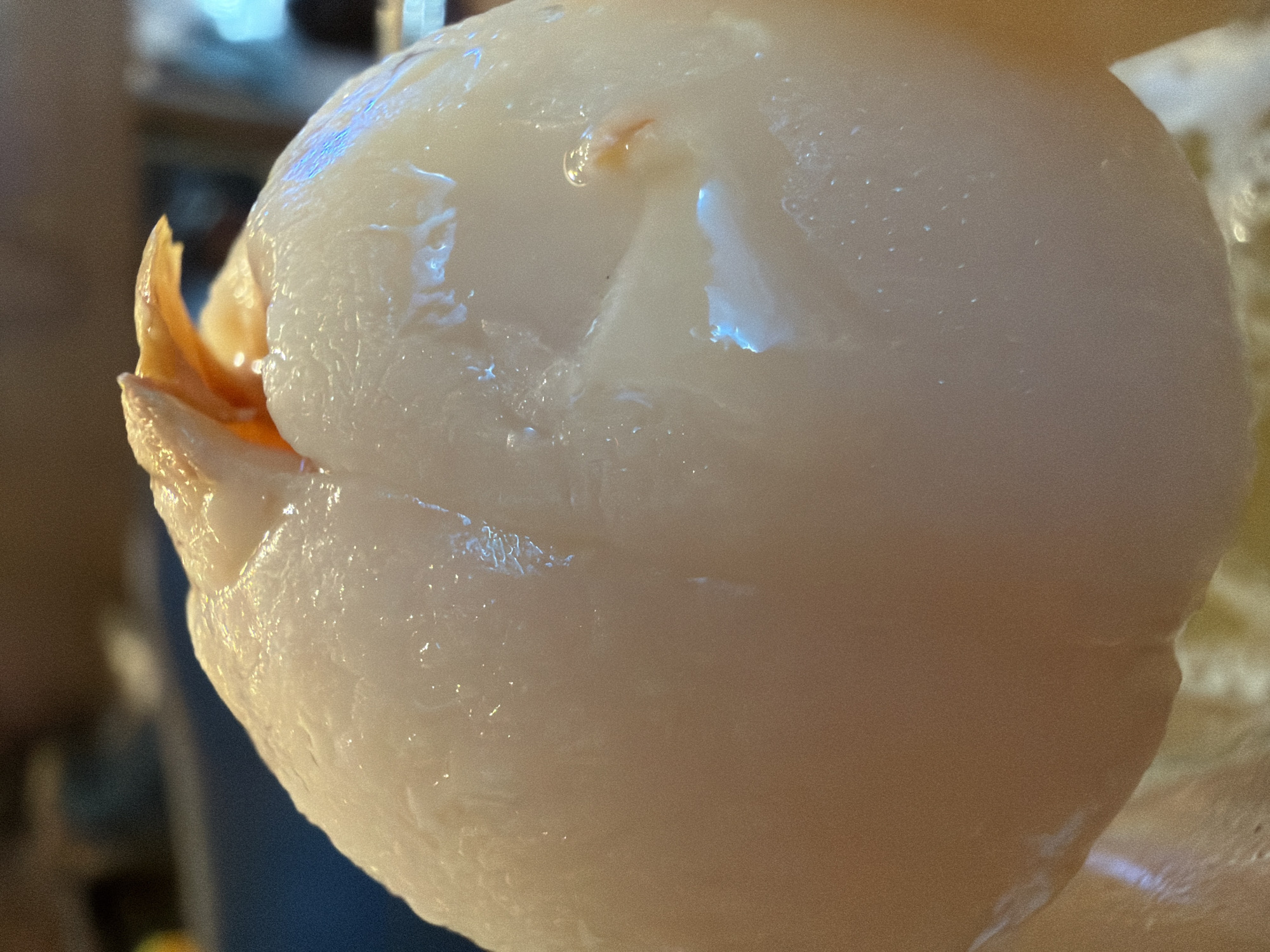 A close-up, macro shot focuses on a peeled lychee fruit. The fruit is smooth and pale, with slight indentations that create shadows and highlight its spherical shape. A small, reddish-orange piece of stem or remnant from the peel is visible at the top of the fruit. The lighting creates reflections and a glossy sheen on the fruit's surface, suggesting it is fresh and possibly wet. The background is blurred, with hints of warm brown tones and a cool blueish area, providing no discernible context about the location within Bac Nha Trang Ward, Vietnam.