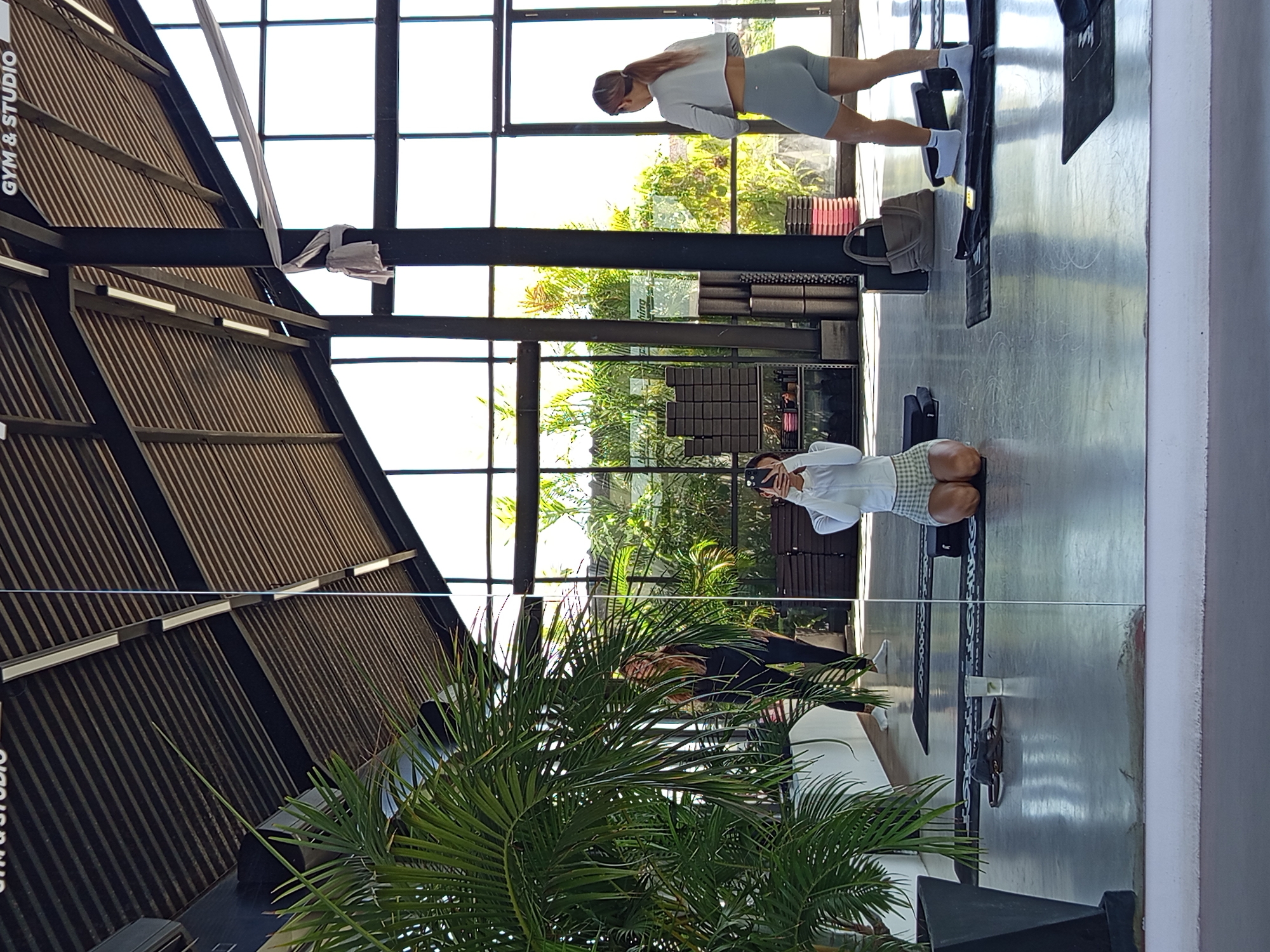 A fitness studio in North Kuta, Indonesia, features an open-air design with large windows overlooking greenery. There are three women present: one doing planks, one kneeling and holding a phone, and a third further in the background. Yoga mats and blocks are visible, indicating a focus on yoga and related fitness activities. A tropical plant, possibly a palm, dominates the foreground, adding to the open-air ambience. The studio structure incorporates dark metal beams and a wooden-slatted roof.