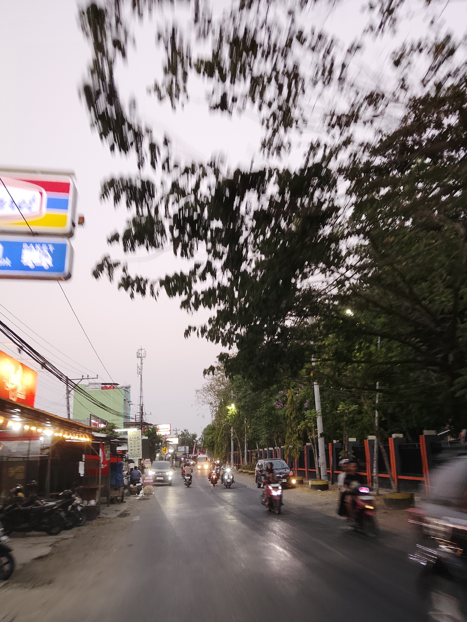 The image captures a street scene in Ngelo, Indonesia, likely at dusk or dawn due to the dim lighting and overcast sky. The perspective appears to be from a moving vehicle, resulting in motion blur across much of the frame. A road with wet or reflective asphalt runs through the center of the image, populated by several motorcycles and a few cars. The traffic consists primarily of motorbikes, some with visible riders, moving in the same direction as the presumed camera. On the left side of the road, a row of low-rise buildings line the street, with some displaying illuminated signs. One sign, partially obscured by blur, features red, yellow, and blue stripes. Further down the street, a cell tower is visible. On the right side, a dense line of trees borders the road, with some foliage appearing in the foreground and background, also showing motion blur. Streetlights are sparsely visible, casting faint glows on the wet road surface. The overall atmosphere suggests a typical urban or suburban street in Indonesia, with active local traffic and commercial activity.