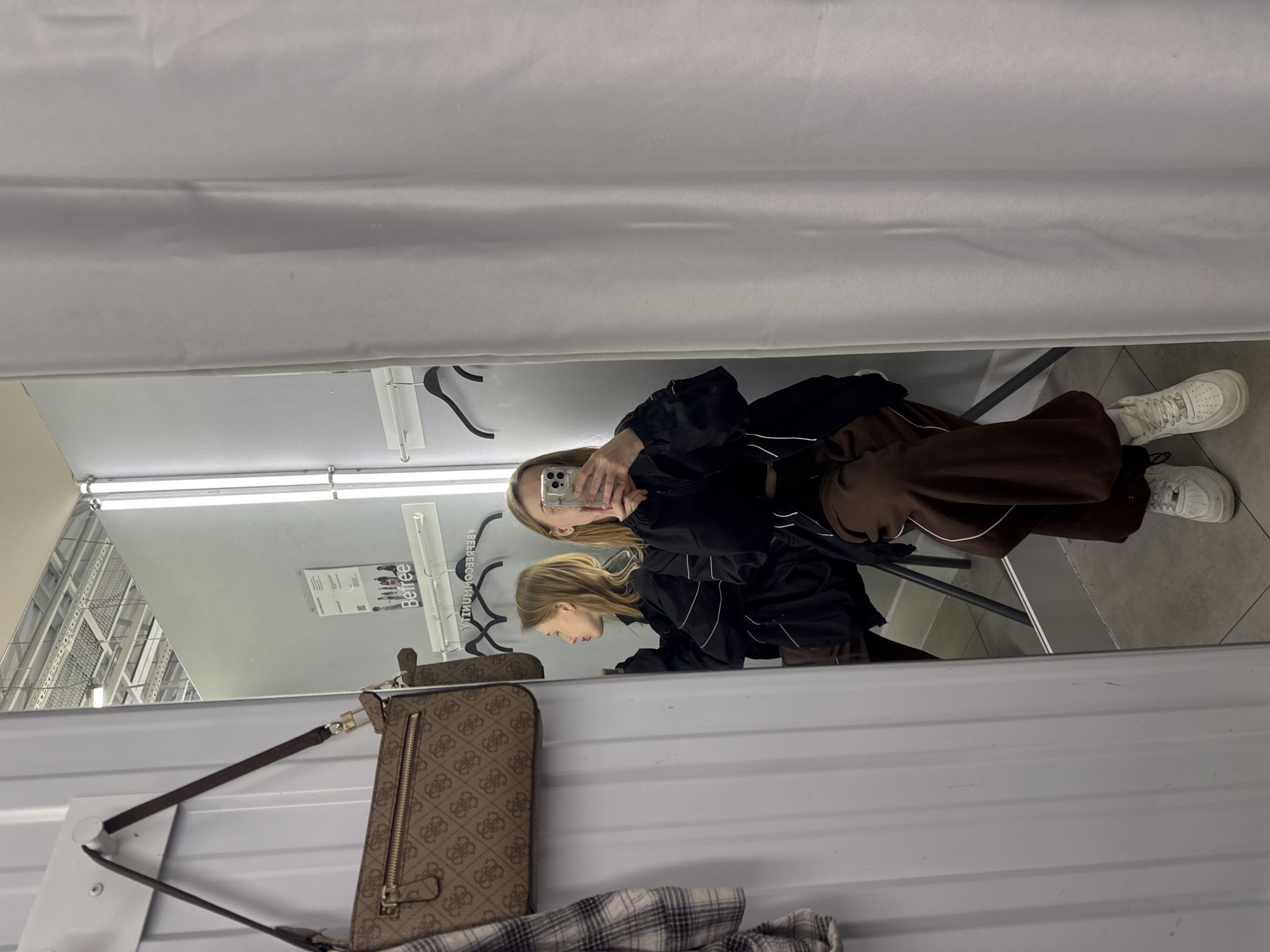 A mirror-reflected image appears to be taken inside a clothing store dressing room in Rostov-on-Don, Russia. A blonde woman in a black jacket and brown pants is taking a photo of herself in the mirror with a smartphone. The floor is tiled, and a chair is visible. A purse hangs on the wall. The store's clothing rack and signage are reflected in the mirror as well. A dressing room curtain is visible at the top of the shot.