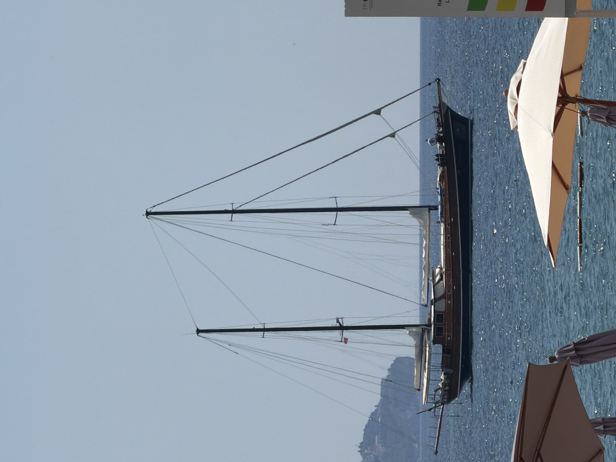 A medium-shot, eye-level, outdoor photograph captures a tall-masted wooden sailing vessel gliding across calm, deep blue water under a clear, pale blue sky. The sailboat, with its multiple sails furled, appears to be at anchor or moving slowly. A few indistinct figures are visible on the deck of the boat. In the background, a hilly, green coastline is dotted with buildings, indicative of a coastal town. In the foreground, two large, beige beach umbrellas are partially visible, with their poles and folded fabric, suggesting a beach setting. A small portion of a white informational sign with colored rectangles is visible on the right side of the frame. The overall scene evokes a serene, leisurely atmosphere. The location is identified as Budva, Montenegro.