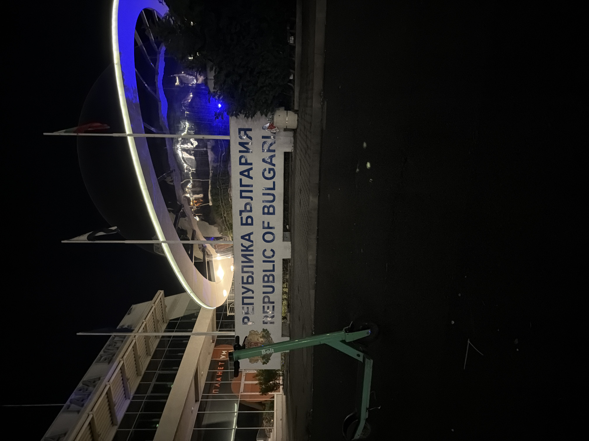 The image shows an outdoor scene at night in Burgas, Bulgaria, featuring a modern architectural structure with a large, illuminated circular element. The structure appears to be part of the Planetarium Burgas, as indicated by visible lettering in Cyrillic on the building's facade. A sign stating "Република България REPUBLIC OF BULGARIA" is present, indicating the location. A Bolt electric scooter is parked on the pavement. Flags are attached to the Planetarium's facade. The scene is illuminated by artificial lights. The area surrounding the building appears dark, suggesting a nighttime setting.