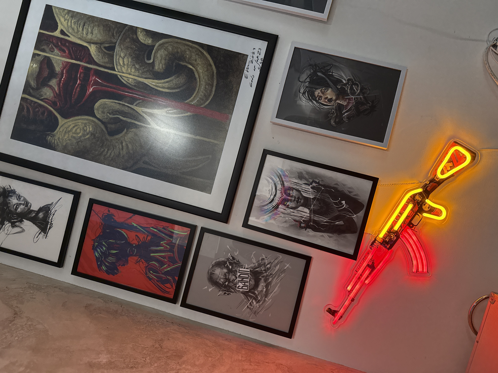 The image depicts a wall adorned with several framed art pieces and a neon light fixture in the shape of an AK-47 assault rifle. There are six framed prints arranged at various angles on a white wall. The largest print, in the upper left, features a dark, textured abstract design. A smaller print to the right of the large one shows a female figure in grayscale. Below these, there are four additional framed prints, each featuring a different grayscale or colored design. The neon AK-47 is mounted on the wall to the right of the prints. The colors of the neon light are red and yellow. The photograph is taken at an angle, suggesting a lower vantage point looking upwards towards the wall. The location is Sofia, Bulgaria.