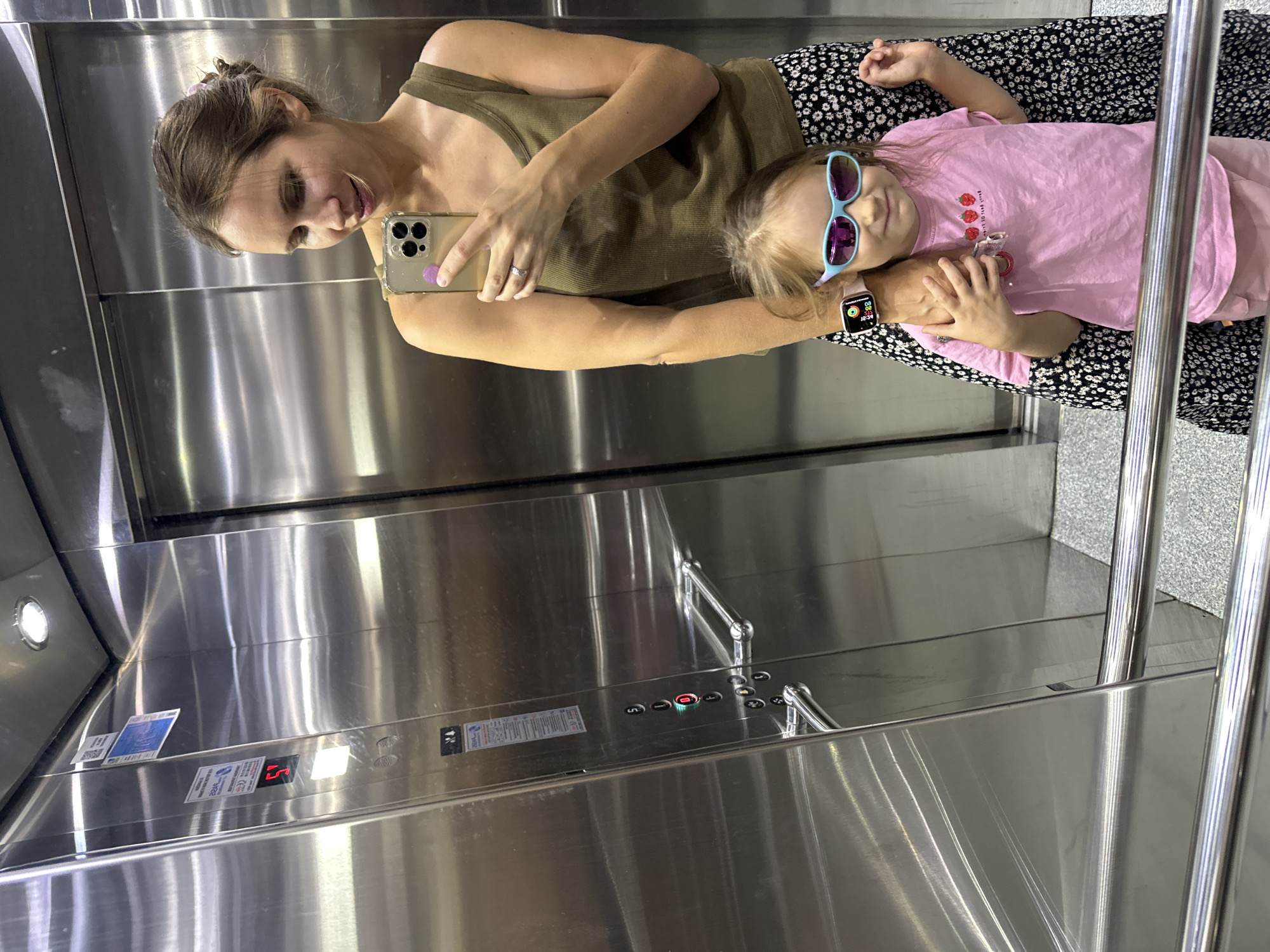 A woman and a young girl are taking a selfie in an elevator. The woman is holding her phone up to take the picture, and the girl is standing in front of her, smiling. The elevator is modern and made of stainless steel. The woman is wearing a green tank top and a floral skirt, and the girl is wearing a pink t-shirt and sunglasses. The girl is also wearing a smartwatch on her left wrist. The elevator appears to be in a building in Fethiye, Turkey. The elevator has a floor indicator displaying "5" and control buttons. There are handrails for passengers. The overall lighting is bright and even.