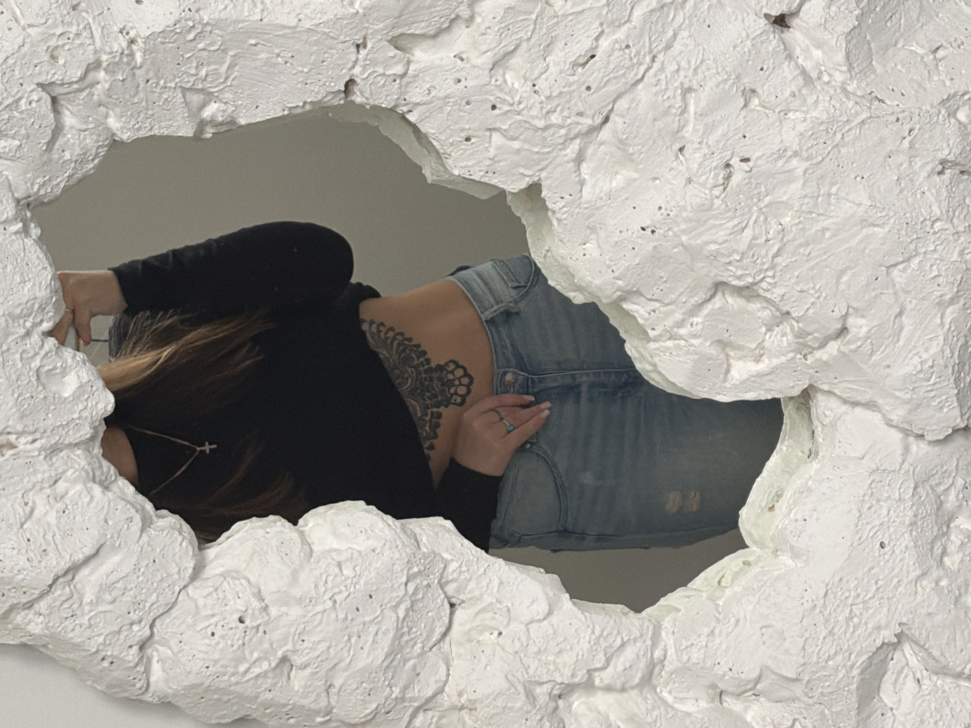 An overhead shot captures a person framed within a ragged, white, heavily textured opening. The individual, appearing to be female, is positioned horizontally, with her back, torso, and part of her head visible through the irregular aperture. She is wearing a long-sleeved black shirt and light blue denim jeans or shorts. A large, intricate black tattoo with a floral or mandala-like design adorns her left upper back and shoulder. Her long, brown hair falls around her head and shoulder. A delicate necklace with a small cross pendant hangs around her neck. On her right hand, a ring with a light-colored, possibly blue, stone is visible.
The white structure that frames the person is rough and uneven, resembling a broken wall or a natural rock formation, with numerous indentations and raised areas. It dominates the foreground and edges of the image, creating a sense of depth and a portal-like view. The background visible through the opening is a plain, light grey surface, likely an indoor wall. The person's posture suggests she is either reclining or interacting with the structure, possibly posing for the photograph. No explicit activity, text, specific location cues, or strong emotions are discernible from the partial view.