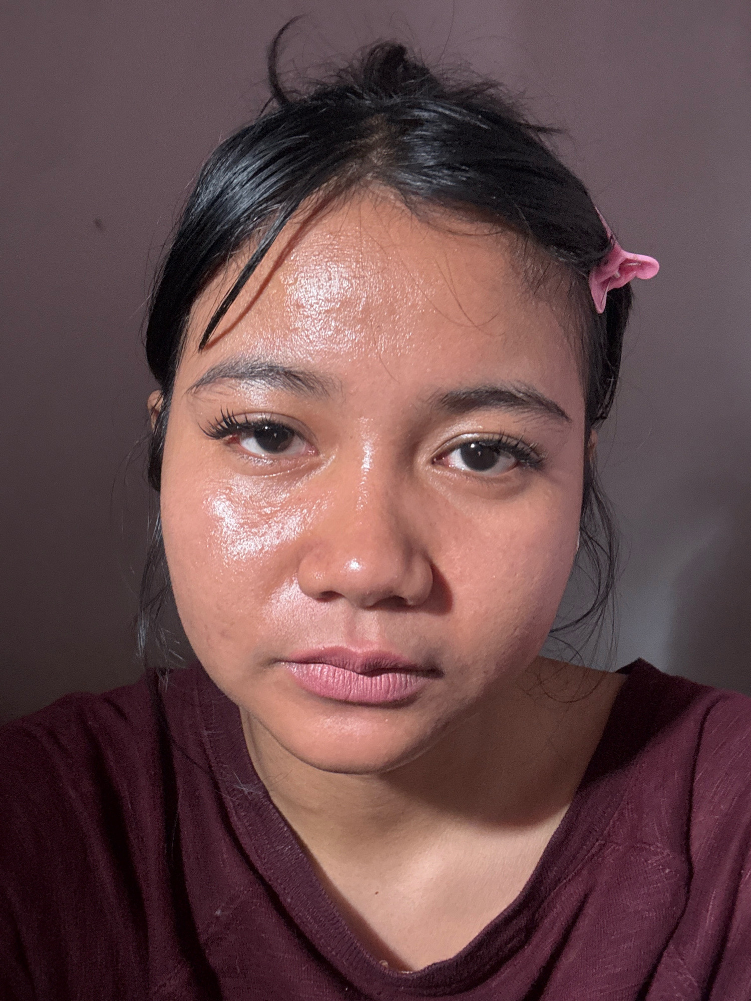 The image shows a close-up, head-and-shoulders portrait of a young woman with dark hair pulled back and secured with a pink clip. She has dark eyes, dark eyebrows, and full lips. Her skin appears dewy and possibly oily, with a slight sheen. She is wearing a dark maroon, collared shirt. The background is a plain, muted purple-gray color. The lighting casts some shadows, particularly on the left side of her face. There are no identifiable landmarks or elements that would suggest a specific location in North Jakarta, Indonesia.