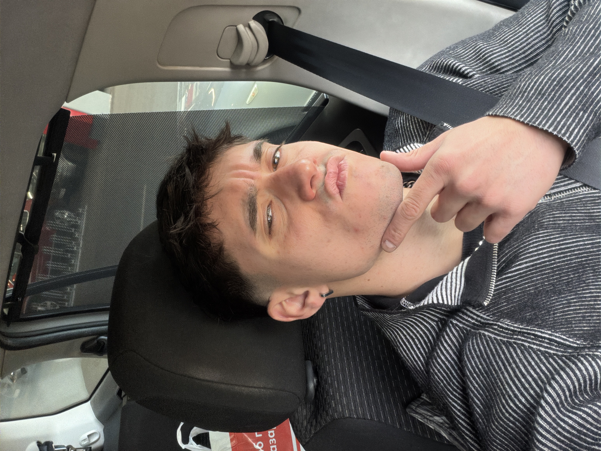 A male individual is seated in the front passenger seat of a vehicle, captured from the chest up. He has short, dark hair with a faded haircut and is wearing a dark gray and white striped long-sleeved top with a zippered collar. A black seatbelt crosses his chest from the upper right. A dark-colored, possibly hexagonal hoop earring is present in his left ear. He looks directly at the camera with a furrowed brow and pursed lips, while his right index finger and thumb touch his chin. There are minor reddish blemishes on the right side of his face. The interior of the car shows a black headrest behind him. To his left, a white plastic bag with red Cyrillic text, reading "магазин" (magazin - shop), is partially visible on the back of the driver's seat. The passenger window is partially covered by a dark mesh sunshade. Through the sunshade, a blurry background consistent with an indoor parking garage or retail parking facility is discernible, showing a reddish structure and dark vertical elements. The presence of Cyrillic text "магазин" indicates the location is within a parking facility in the city of Sofia, Bulgaria.
