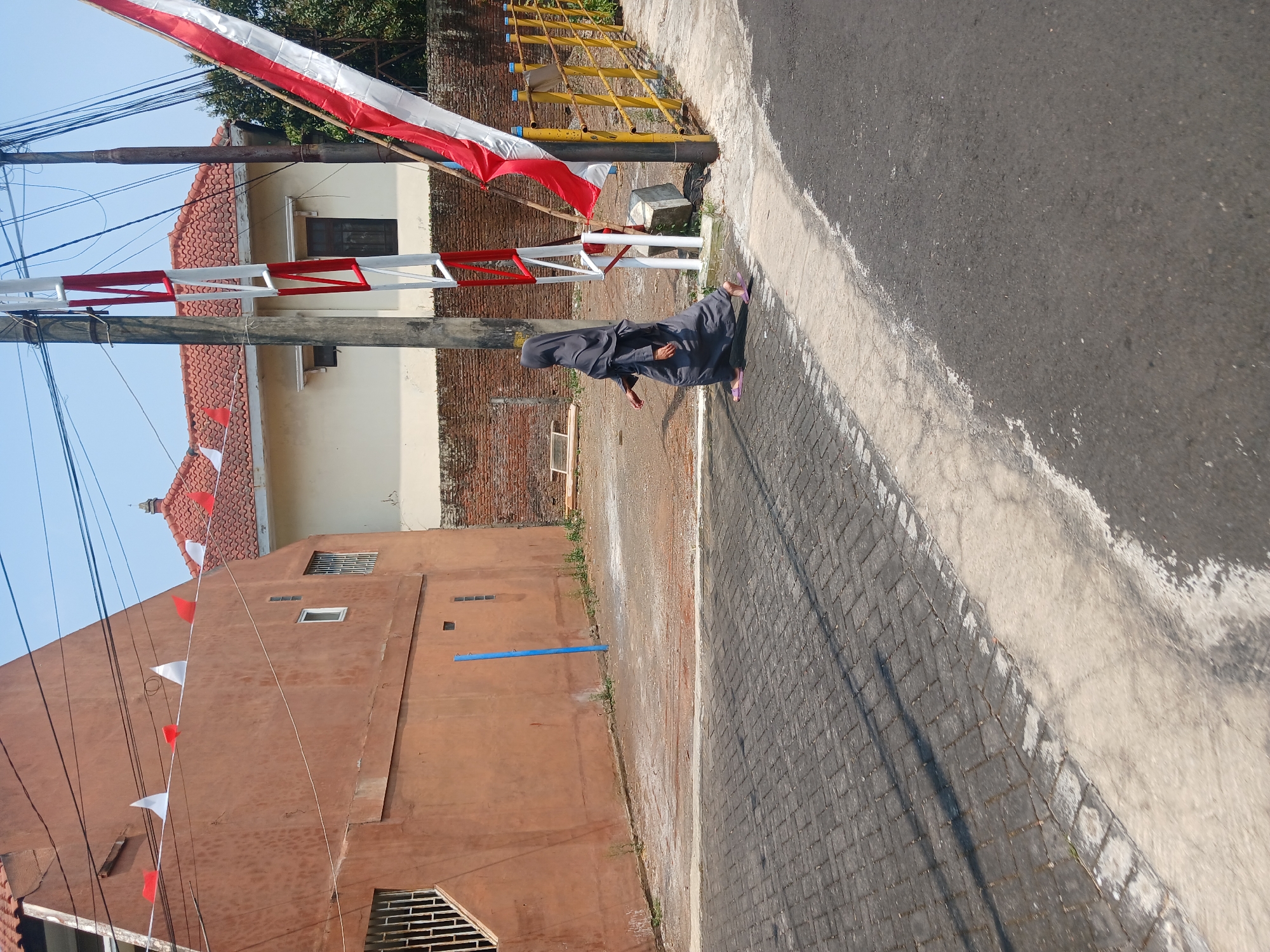 In Watugede, Indonesia, a person wearing a grey hijab and dress walks across a paved street. The street has a border of interlocking grey pavers next to a strip of concrete and then asphalt. To the right of the street is a yellow metal fence and a red and white striped barrier pole with a large red and white flag attached. Behind the fence are trees and a brick wall. In the background, there are buildings with terracotta tile roofs and orange and cream-colored walls. Red and white triangular flags are strung along the top of the orange building. A clear blue sky is visible above.