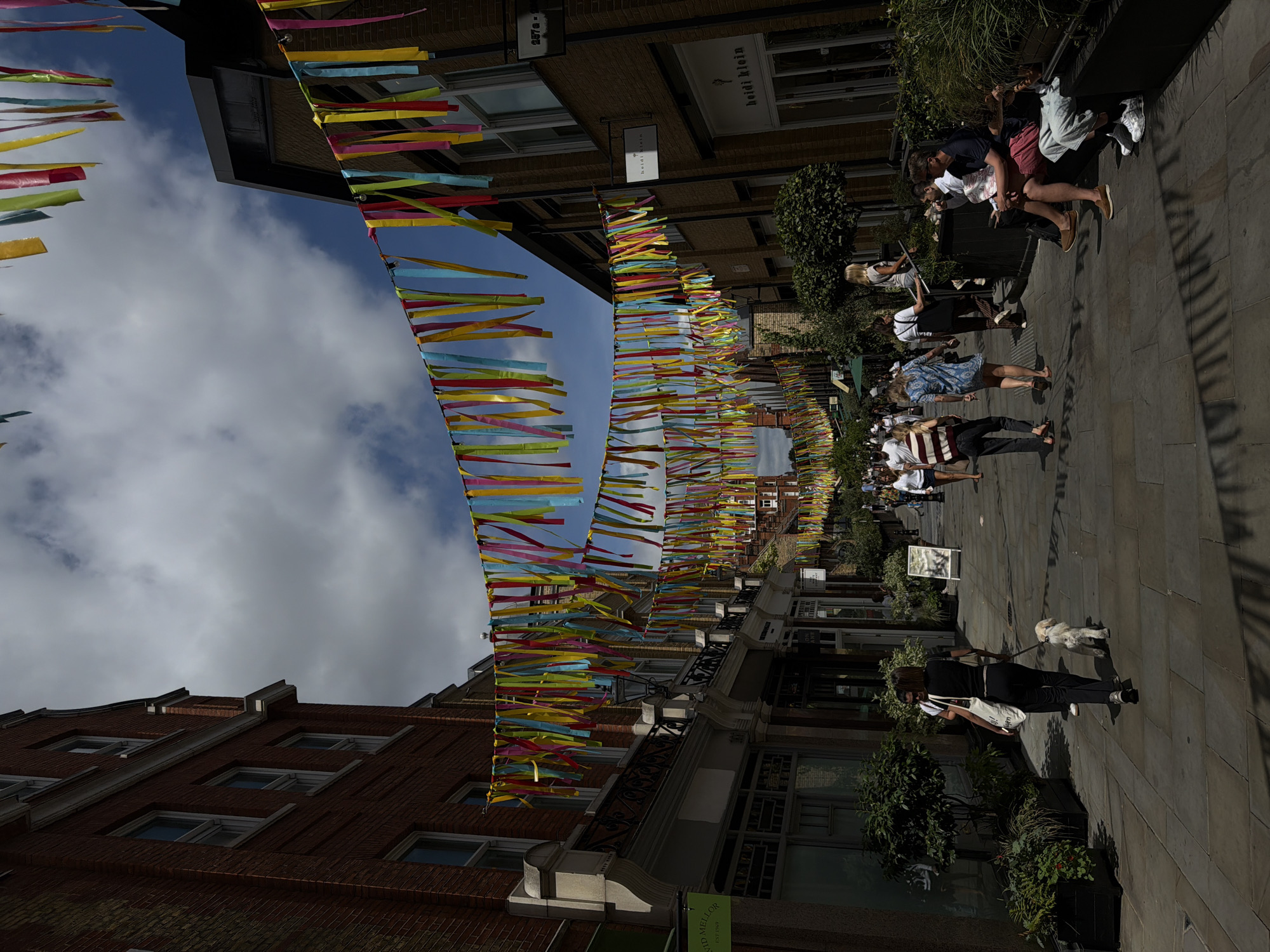 The image depicts a vibrant street scene in the heart of London, United Kingdom. Colorful streamers are strung across the street, adding a festive atmosphere to the urban environment. People are walking along the pedestrianized street, some with their dogs, while others are seated on benches, enjoying the sunny day. The architecture suggests a historic district, with buildings made of brick and stone. Signs for shops like "Heidi Klein" are visible, indicating a commercial area. The scene is bathed in sunlight, casting shadows on the pavement and highlighting the textures of the buildings and the foliage.