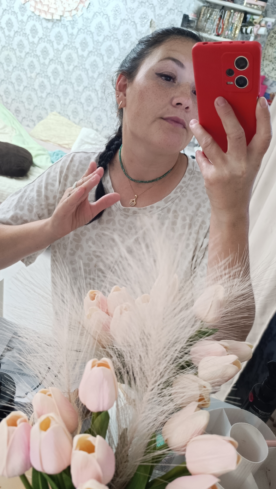 A woman is taking a selfie in what appears to be a bedroom. She is holding a red smartphone in her right hand, angled to capture her reflection. She has dark hair pulled back into a braid and is wearing a light-colored, patterned t-shirt and a necklace with green beads and a gold pendant. Her left hand is raised and positioned near her face, touching her braid. In the foreground, a bouquet of light pink tulips and fluffy white plumes is visible, partially obscuring the lower part of the woman and the immediate surroundings. The background shows a patterned wallpaper, a bed with linens, and shelves filled with various items, suggesting a personal space. The overall lighting is soft, indicating an indoor setting, likely during the daytime. There are no specific landmarks or indicators to place the image in Chaikovsky, Russia, beyond the provided prompt.