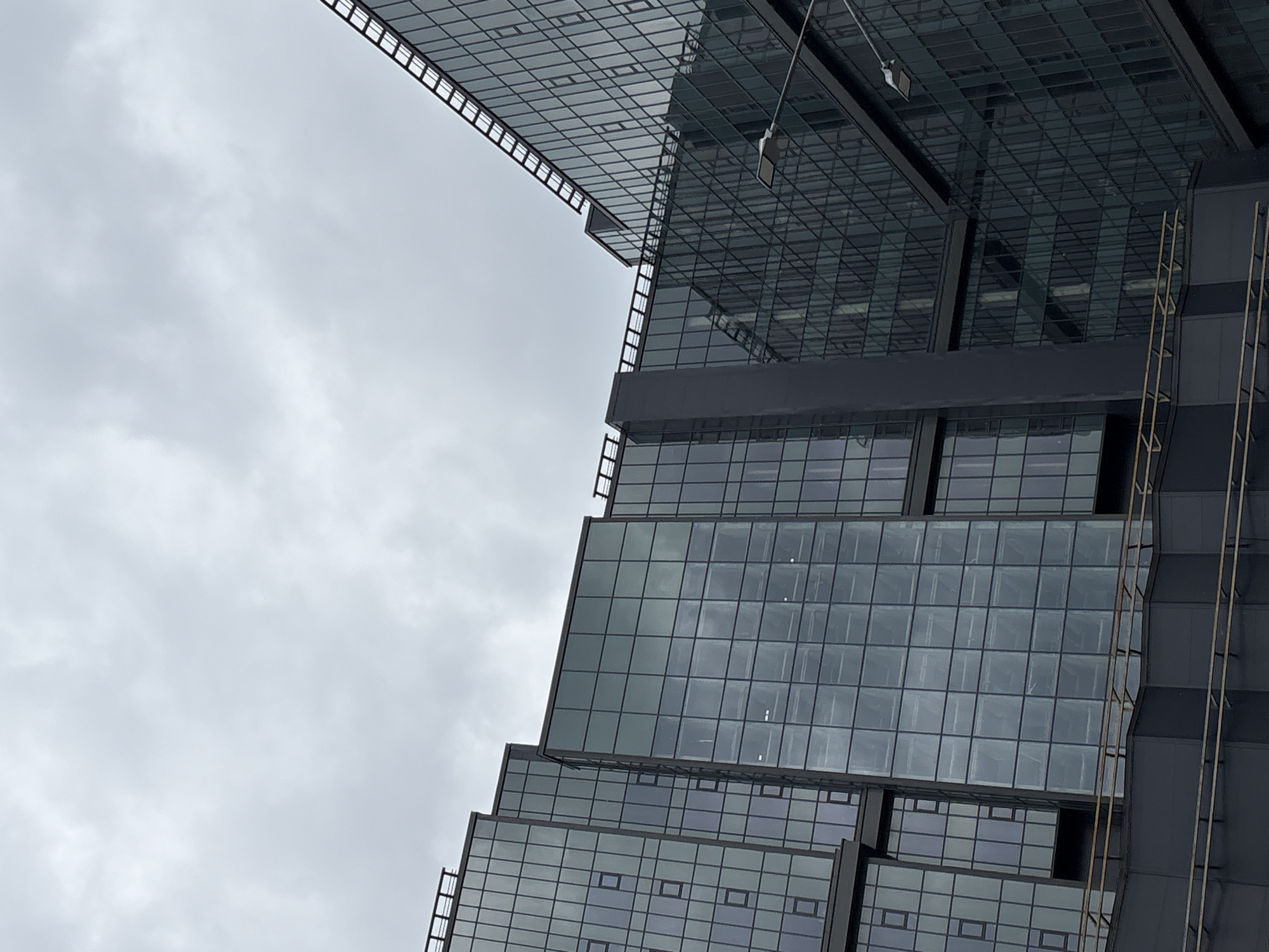 The image shows a close-up of a modern, multi-story glass-and-steel building against an overcast sky. The building features a grid-like facade with numerous windows. Several sections of the building are visible, appearing to be connected at different angles and heights. The sky is uniformly gray and cloudy, with no visible sun or distinct cloud formations. The overall impression is one of an urban environment, likely a business district. The image does not contain any people or identifiable landmarks to pinpoint a specific city. The building's architectural style suggests it could be a contemporary office complex or residential tower.