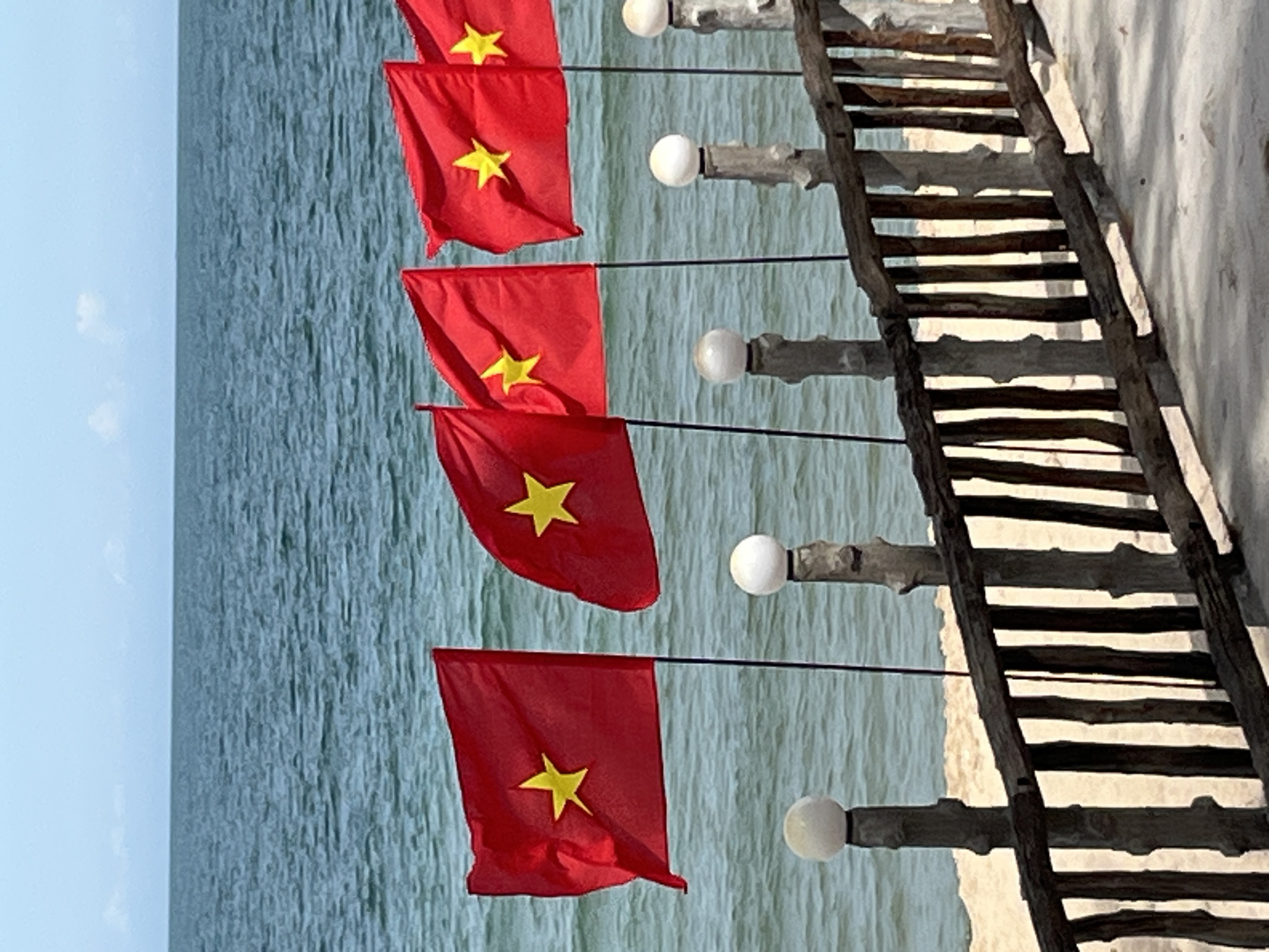 Five red flags with a yellow star in the center, identified as Vietnamese flags, are displayed on flagpoles. These flags are positioned in front of a body of water, likely the sea, under a clear sky with a few faint clouds. A wooden railing runs horizontally across the lower part of the image, supported by rough-hewn posts topped with spherical white lights. Beyond the railing, a sandy area is visible. The scene is likely a waterfront location in Mũi Né, Vietnam, possibly a resort or public area overlooking the ocean. There are no people visible in the image.