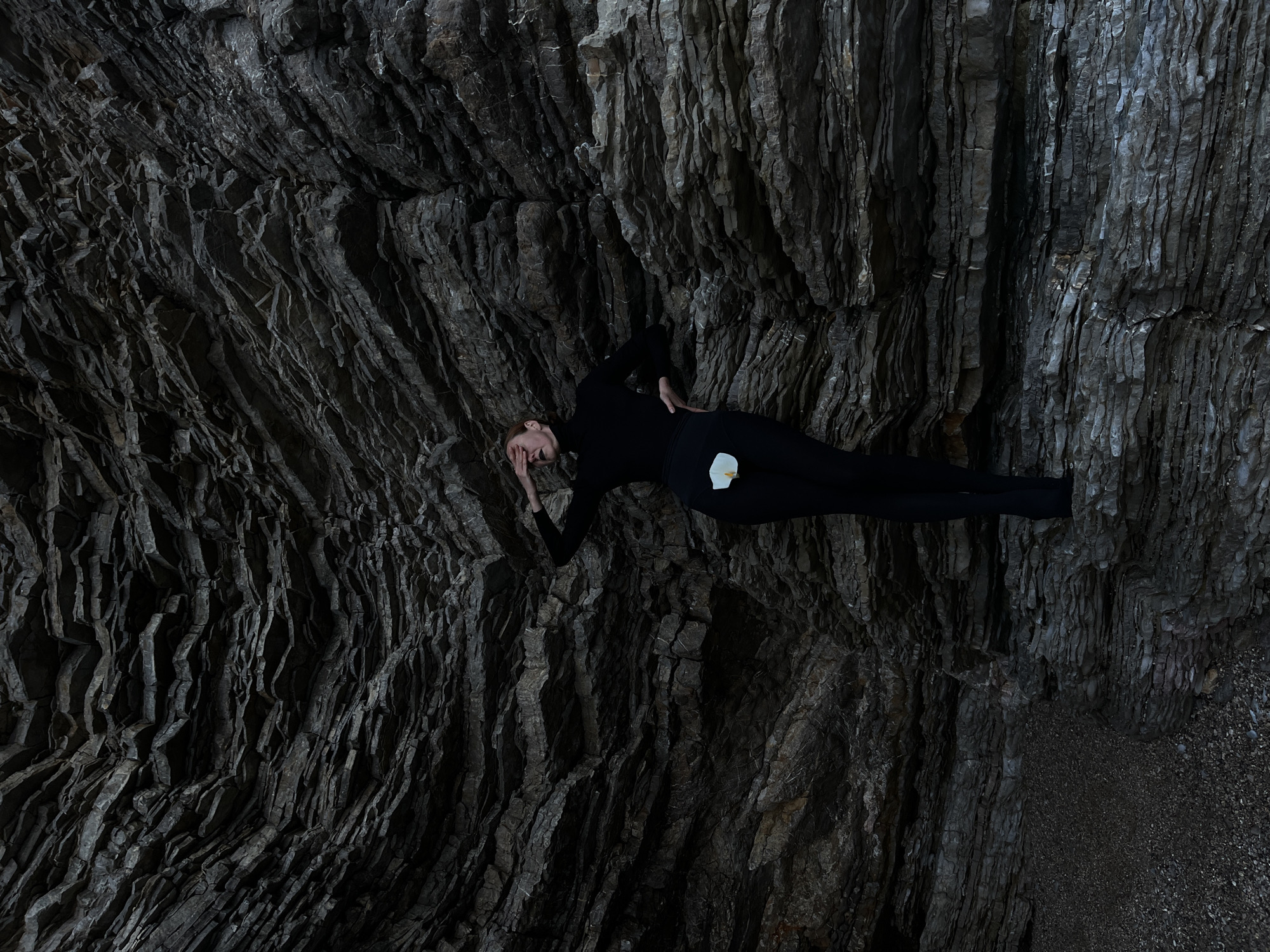 A long shot shows a slender person lying on a rocky outcrop. The person is wearing a black turtleneck and black leggings, and has pale skin. A single white calla lily lies on their hip. Their face is obscured by their hand and arm. The rock has many horizontal layers. The image appears to be taken at a high angle. The location may be the cliffs along the coast of Budva, Montenegro.