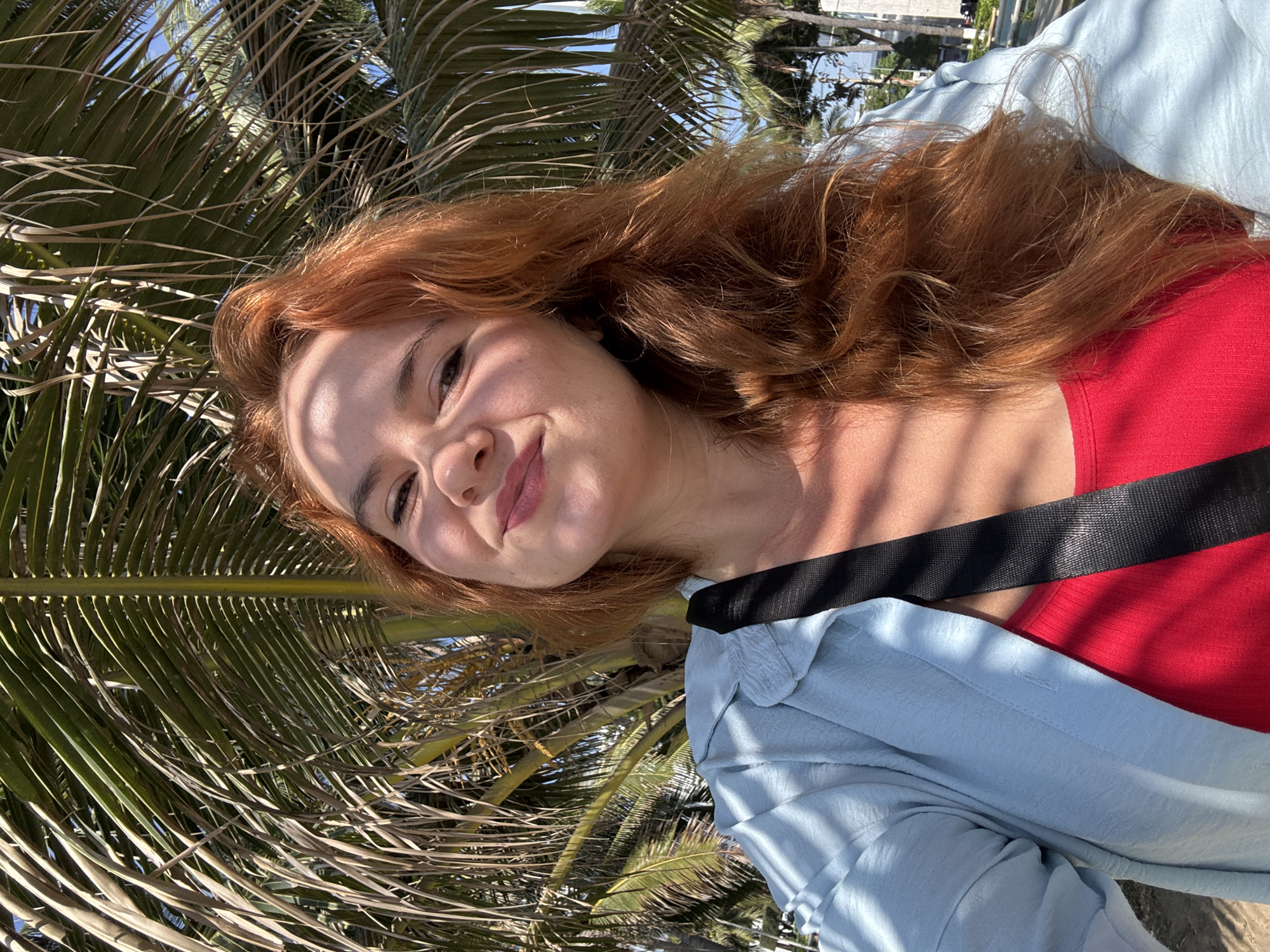 A red-haired woman with fair skin and a light dusting of freckles smiles at the camera. She is wearing a red tank top and a light blue button-down shirt. A black strap is visible across her chest. The background consists of palm fronds with dappled sunlight. The overall impression is of a sunny day, possibly in a tropical location.