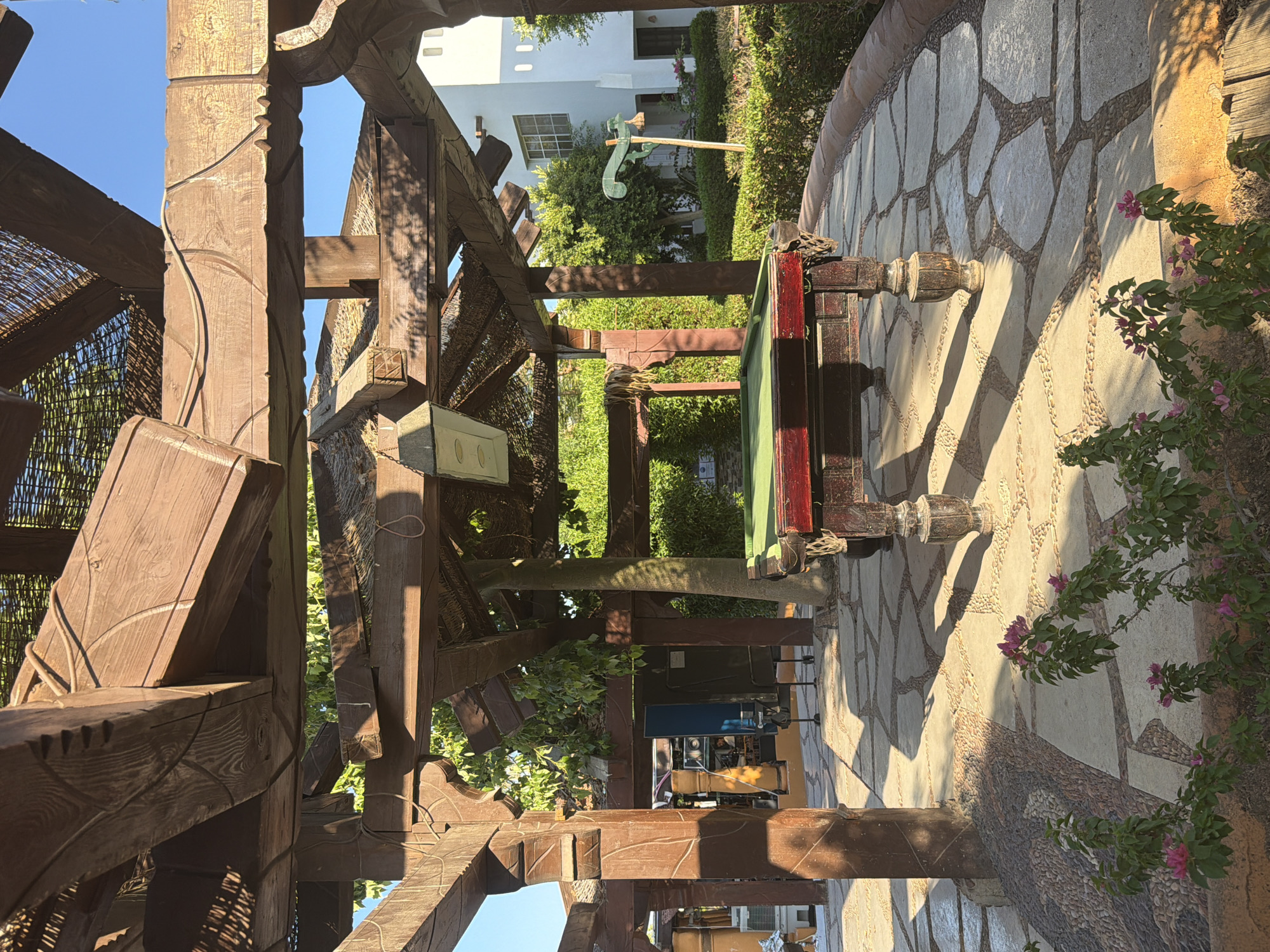 The image depicts an outdoor setting under a rustic wooden pergola, likely a resort or hotel area in Sharm El Sheikh, Egypt. The main focus is a pool table with a green felt surface and a dark red frame, positioned centrally. The table has ornately carved wooden legs. The ground beneath is paved with irregular flagstones interspersed with smaller rounded stones, creating a mosaic-like pattern. Sunlight casts distinct shadows across the paving.
In the foreground, there are flowering plants with pink blossoms and green leaves. Behind the pool table, a large tree trunk is visible, integrated into the structure of the pergola. Further back, lush green foliage and bushes form a backdrop, with a white building with windows visible in the upper right portion of the frame. The sky is clear and blue. There are no people directly visible in the image, but a glimpse of a person is visible through a gap in the pergola structure in the middle-left background, near some equipment. The overall atmosphere suggests a leisure area designed for relaxation and recreation in a warm climate.