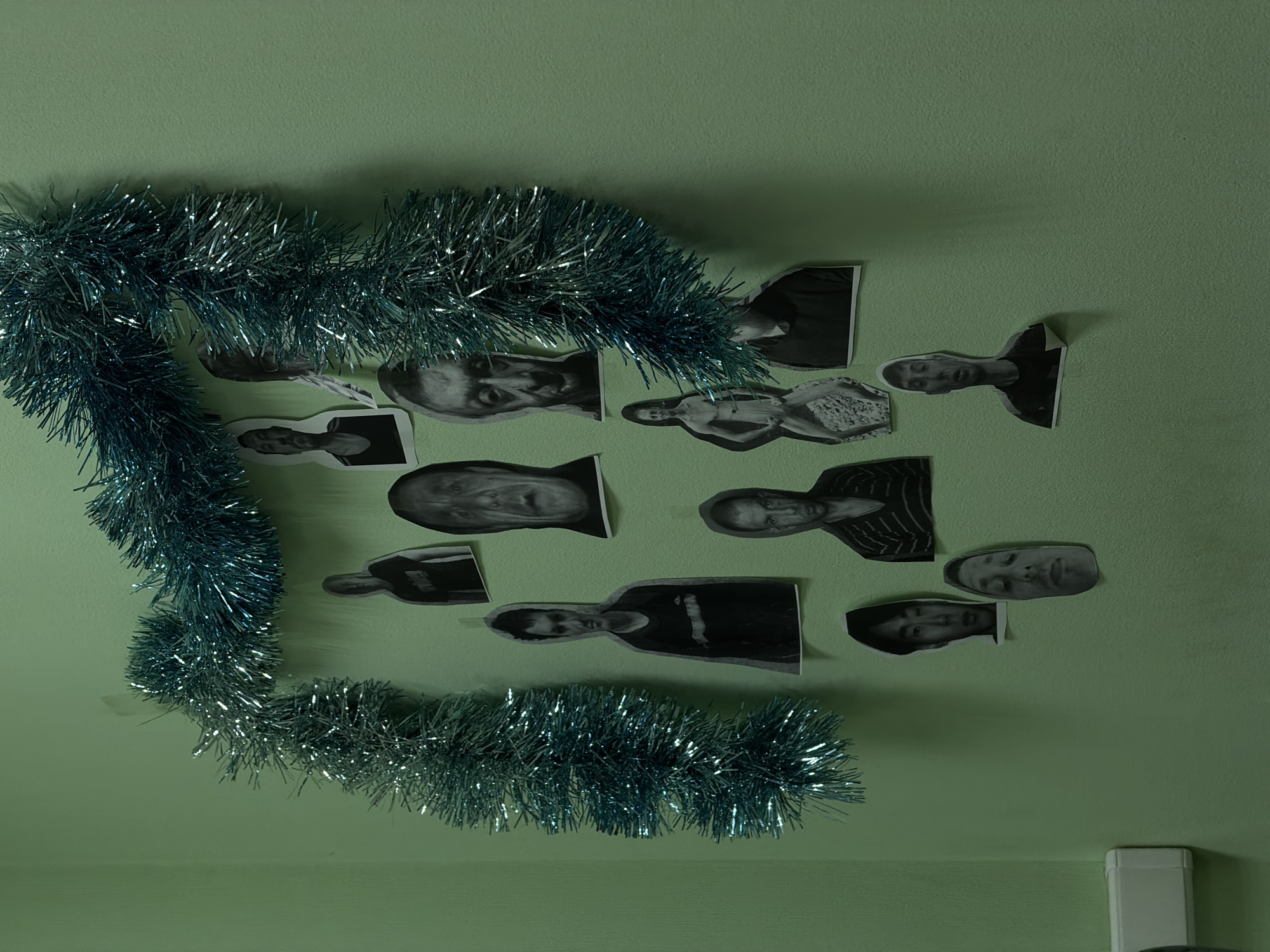 The image shows a light green wall decorated with silver tinsel and black and white cut-out photos of people. The tinsel is arranged in a loose "L" shape. The photos are clustered around the tinsel and seem to be portrait shots. The location is presumed to be in Chaikovsky, Russia, based on the prompt's instruction. There is a white electrical outlet cover visible in the lower right corner of the image.