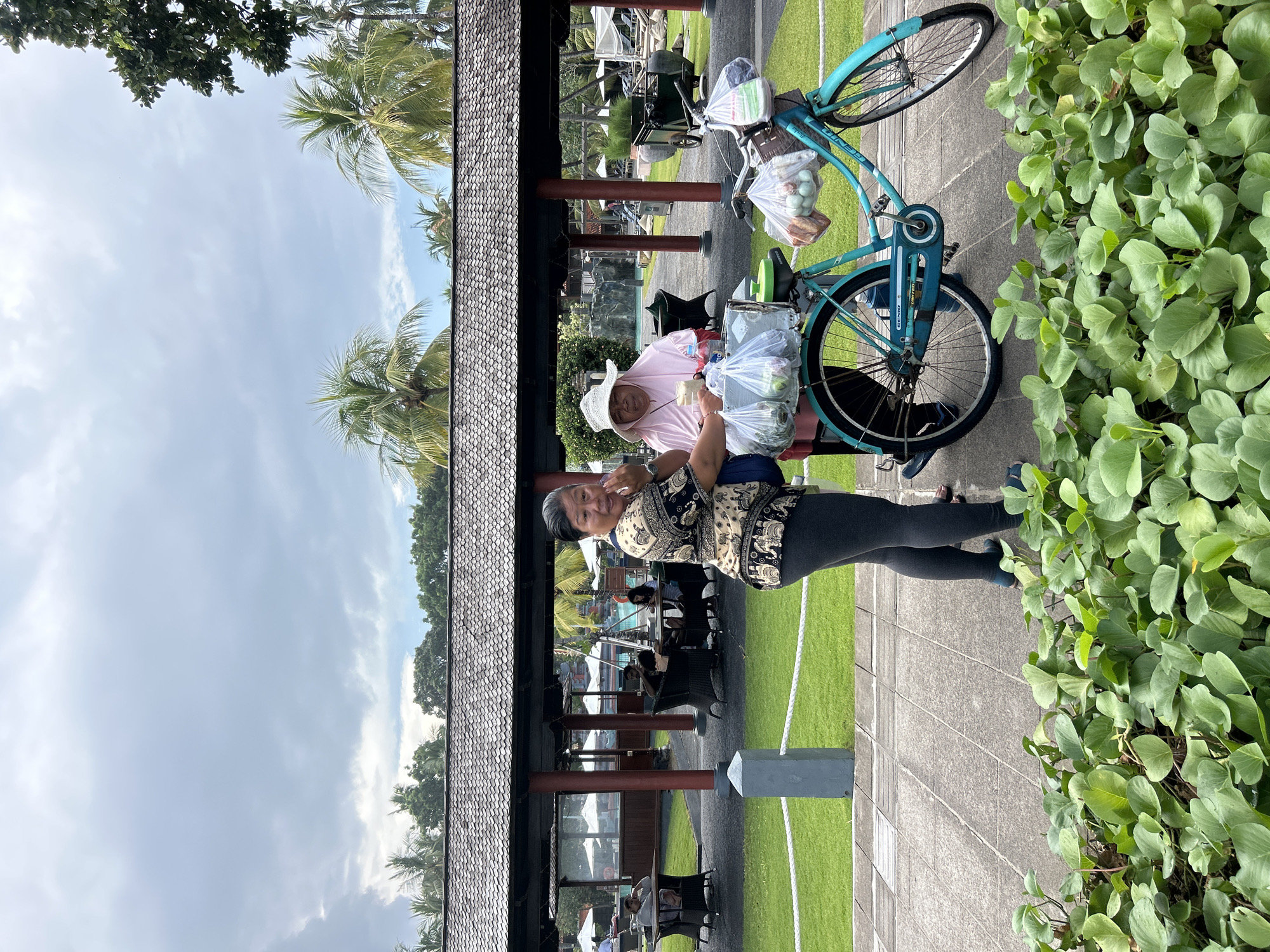 Two women stand near a turquoise bicycle loaded with plastic bags of produce. The woman on the left, wearing a black and beige patterned shirt and dark grey leggings, looks towards the camera. The woman on the right, wearing a pink shirt and a white sun hat, is interacting with the woman on the left and holding a clear plastic cup. The bicycle has bags of what appear to be fruits and vegetables attached to the front and rear. In the background, a covered outdoor seating area with tables and chairs is visible, with people seated at some of the tables. Palm trees and a cloudy sky form the backdrop. The foreground is filled with lush green ground cover plants. The scene appears to be in Kuta, Indonesia, likely at a resort or public area.