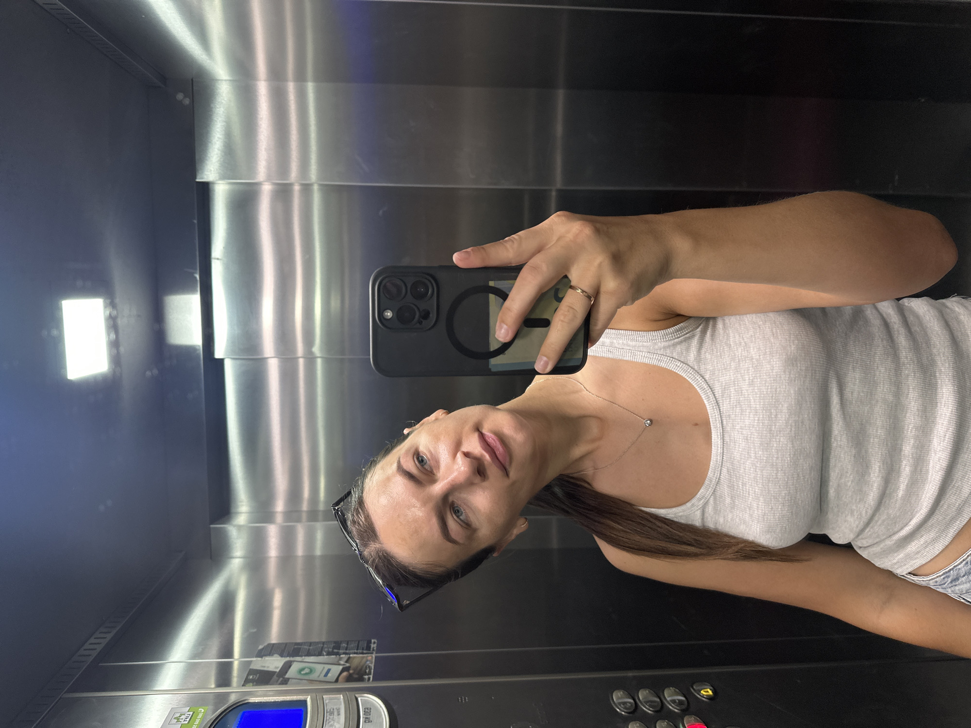 The image shows a woman taking a selfie in an elevator with brushed metal walls and a bright overhead light. She is wearing a grey ribbed tank top, denim shorts, sunglasses on her head, and a delicate necklace with a single pendant. She is also wearing a thin gold ring on her left hand, which is holding her black smartphone. The phone has a circular cutout on the back, and a yellow bill is visible behind it. A small portion of elevator buttons, including a red illuminated button, are visible on the left side of the image. The elevator appears to be in a modern building, possibly a hotel or residential complex in the city of Budva, Montenegro, given the context.