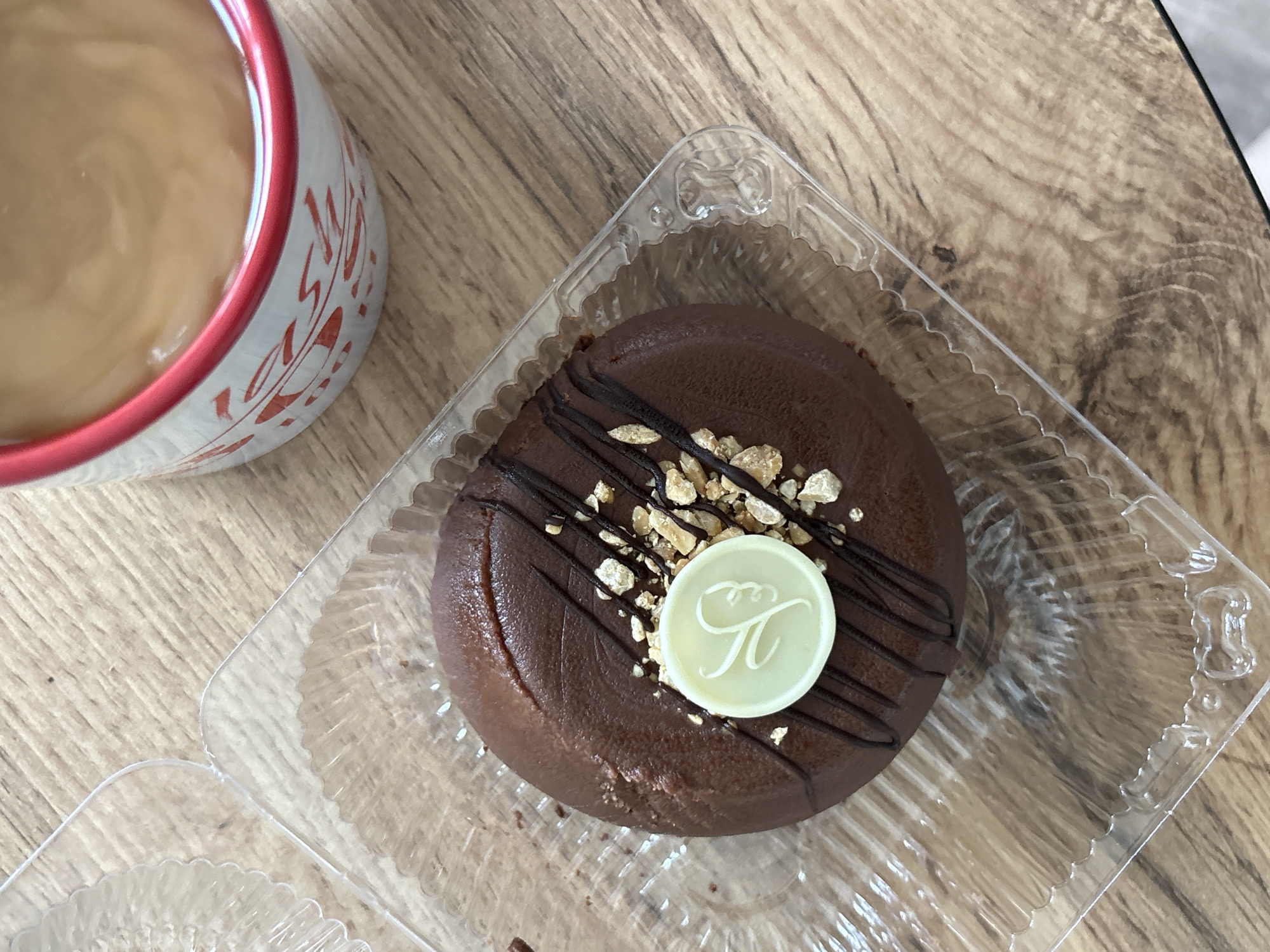 A chocolate pastry with a white decorative emblem and chopped nuts sits in a clear plastic container on a wooden table. To the right of the pastry, a white mug with a red rim, decorated with red script, contains a light brown beverage. The scene is viewed from directly above. The location is not determinable from the image.