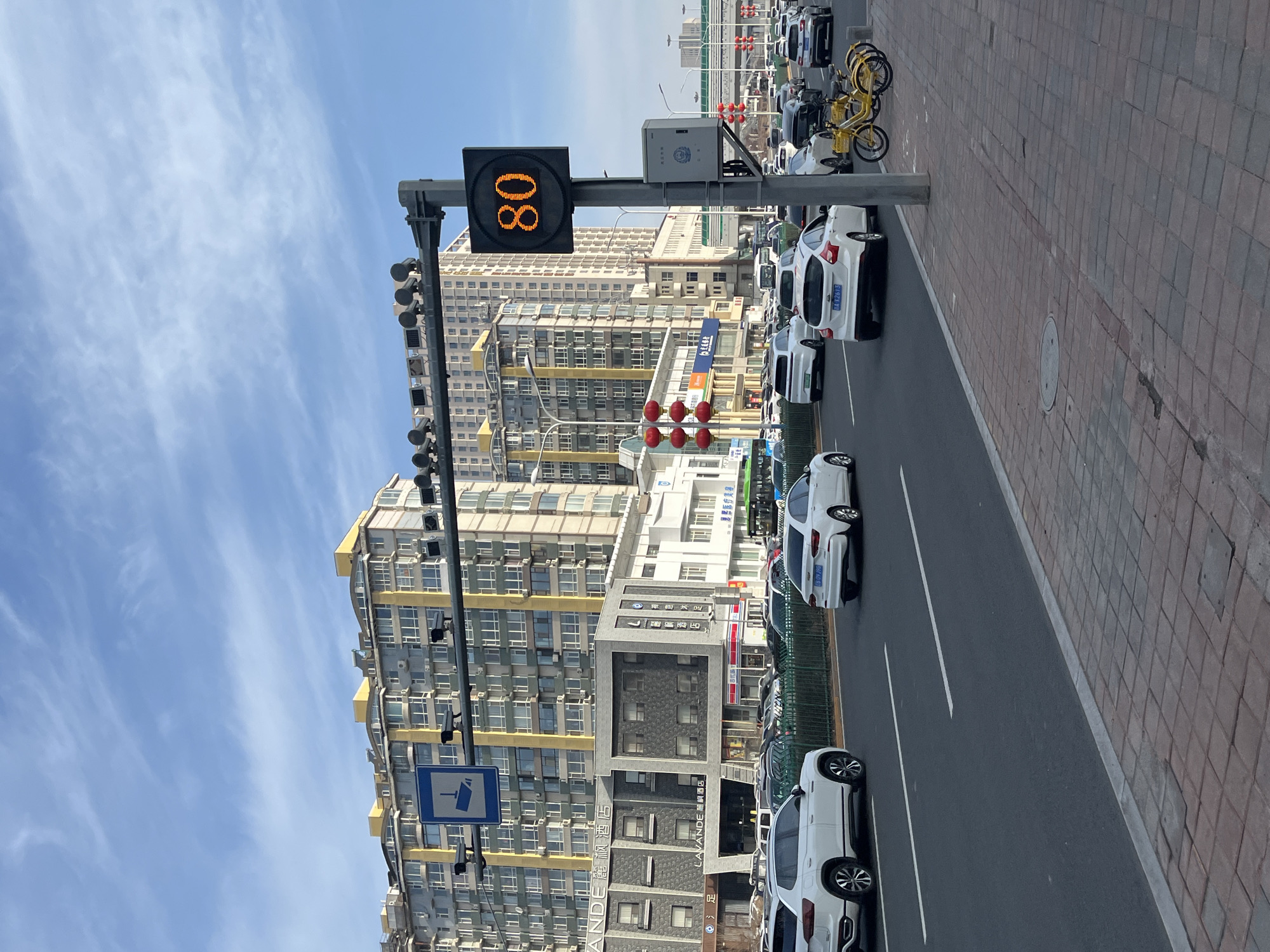 The image shows a street scene in Changchun City, China. On the left, there is a digital speed limit sign displaying "80" atop a traffic pole with multiple surveillance cameras. Behind the pole, there are tall buildings, primarily residential, with a mix of modern and older architectural styles. A Lavande Hotel is also visible among the buildings. On the right, there is a row of parked white cars along the roadside. Further back, there are some yellow bicycles and more vehicles. The sky is blue with scattered clouds. The sidewalk is paved with reddish-brown bricks.