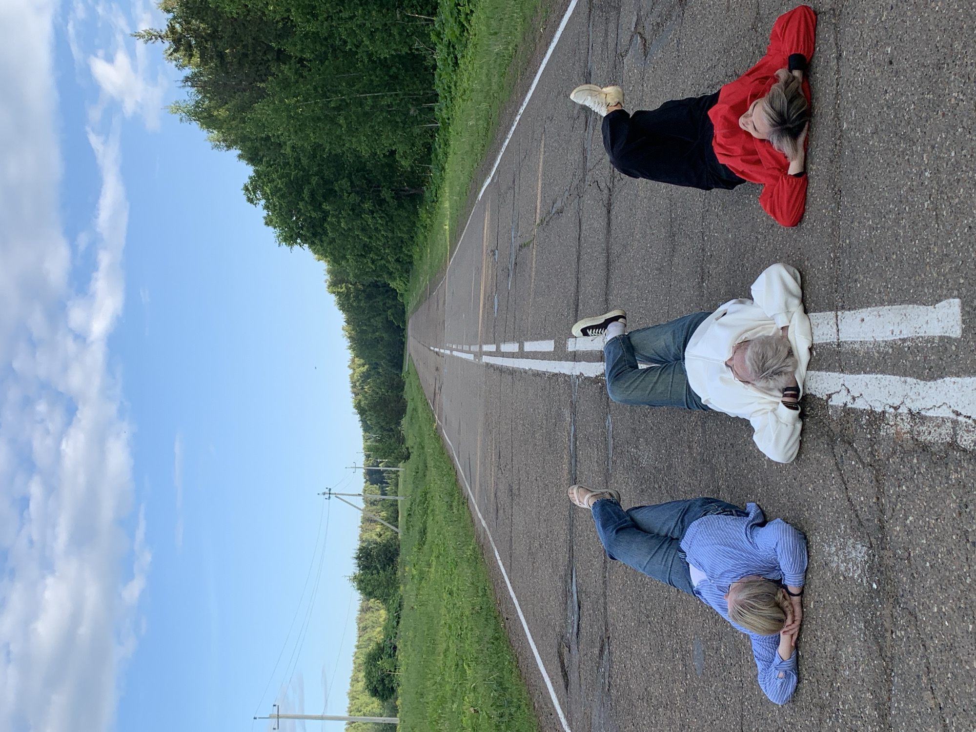 An image, initially oriented vertically with the sky to the left, depicts three individuals lying horizontally on their backs on a paved, rural road, looking up at a bright blue sky. This scene takes place in what appears to be Bol'shoi Bukor, Russia.
The road itself is asphalt, showing signs of wear and cracks, and stretches into the distance, converging towards a vanishing point. It is marked by a continuous white line along its edge and a dashed white line in the center. Dense green trees and forest line both sides of the road, creating a natural, secluded corridor. Utility poles are visible along the right side of the road in the mid-ground.
Three people, appearing to be young adults, are spaced out along the road's surface. The person closest to the foreground wears a blue and white striped long-sleeved shirt and blue jeans, with light-colored hair. Their hands are clasped near their midsection. The second person, further along the road, is dressed in a white hooded sweatshirt and blue jeans, also with light hair, and rests their hands near their head. The third person, positioned furthest from the camera, wears a red and black top with black pants, and has light hair, with their hands behind their head. All three are lying still, seemingly relaxed, and gazing upwards. The time of day appears to be daytime, with a clear blue sky dotted with scattered white clouds, suggesting pleasant weather. There is no visible text in the image.