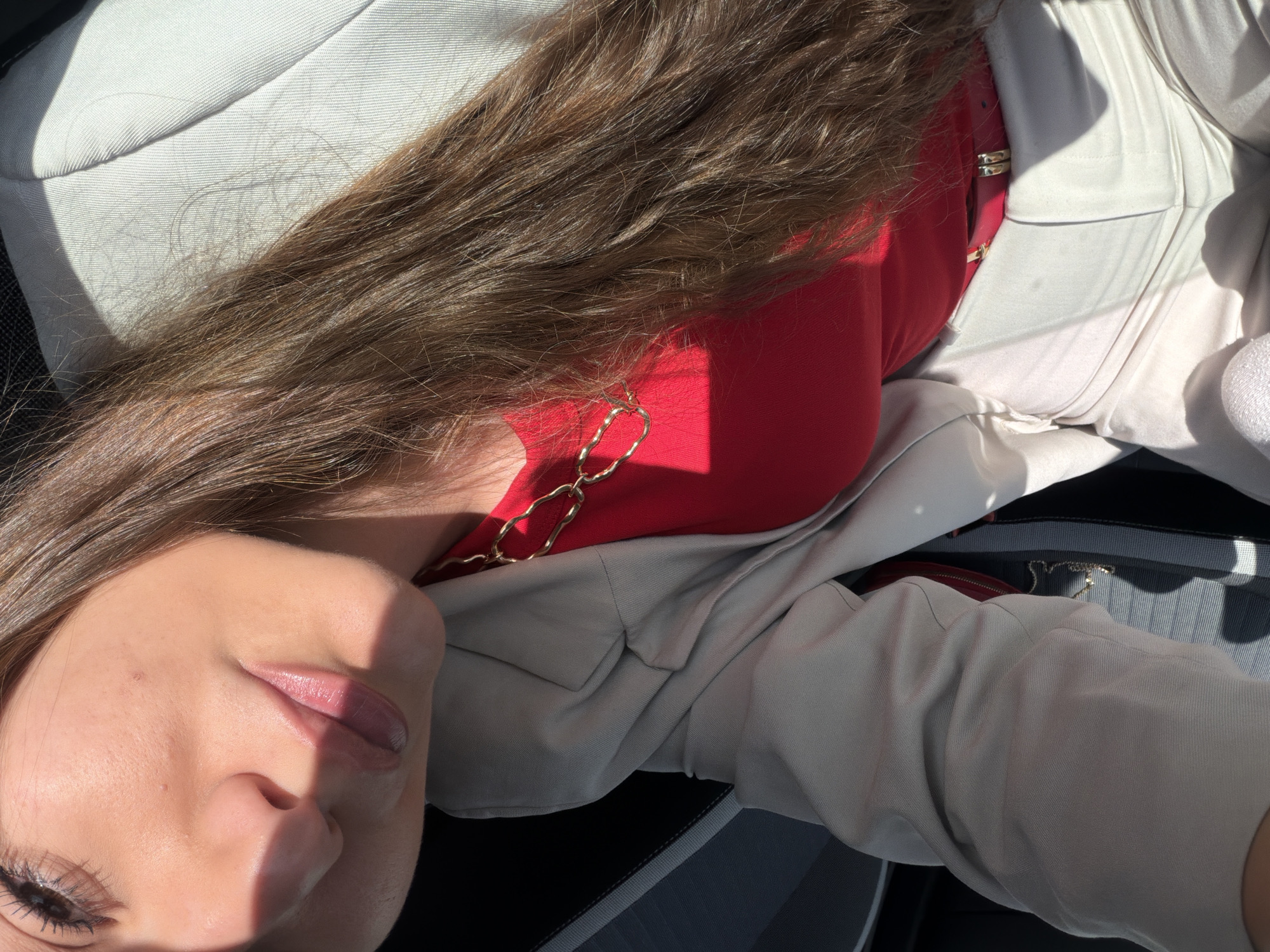 A female individual is partially depicted in a close-up, high-angle view, seated within the interior of a vehicle. Her face, upper torso, and lap are visible. She has fair skin, visible brown hair with lighter highlights, and appears to be wearing mascara and lip gloss. Her attire consists of a red top, a light beige or off-white blazer/jacket, and matching light beige or off-white pants. A gold-colored chain necklace with irregular links is present on her chest, and a gold-colored belt buckle is partially visible at her waist. Strong, direct sunlight illuminates the left side of her face and upper body, creating a sharp shadow line across her nose and mouth. Loose strands of brown hair are visible on her pants. A red object, possibly a small bag or wallet, is partially discernible near her right hip. The vehicle interior shows a dark grey ribbed seat with light-colored stitching. The precise location within Sofia, Bulgaria, is not discernible from the image.