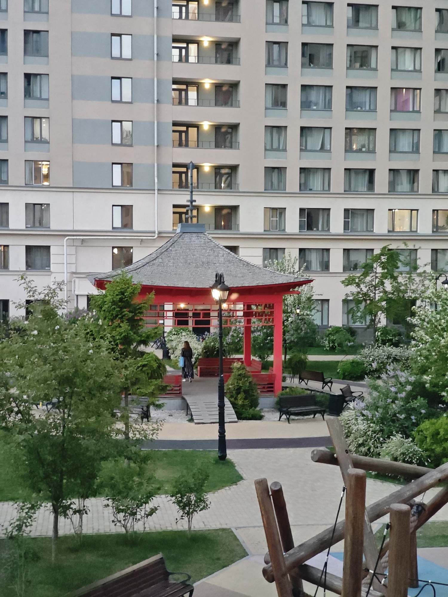 The image captures a cityscape in Yekaterinburg, Russia, featuring a Japanese-style gazebo amidst a landscaped park. The gazebo is red with a grey roof, and a woman in a black and white striped dress is walking towards it. The park includes trees, shrubs, benches, and a playground structure made of logs. In the background, a tall apartment building with multiple windows is visible. The scene suggests an evening setting with the building lights turned on.