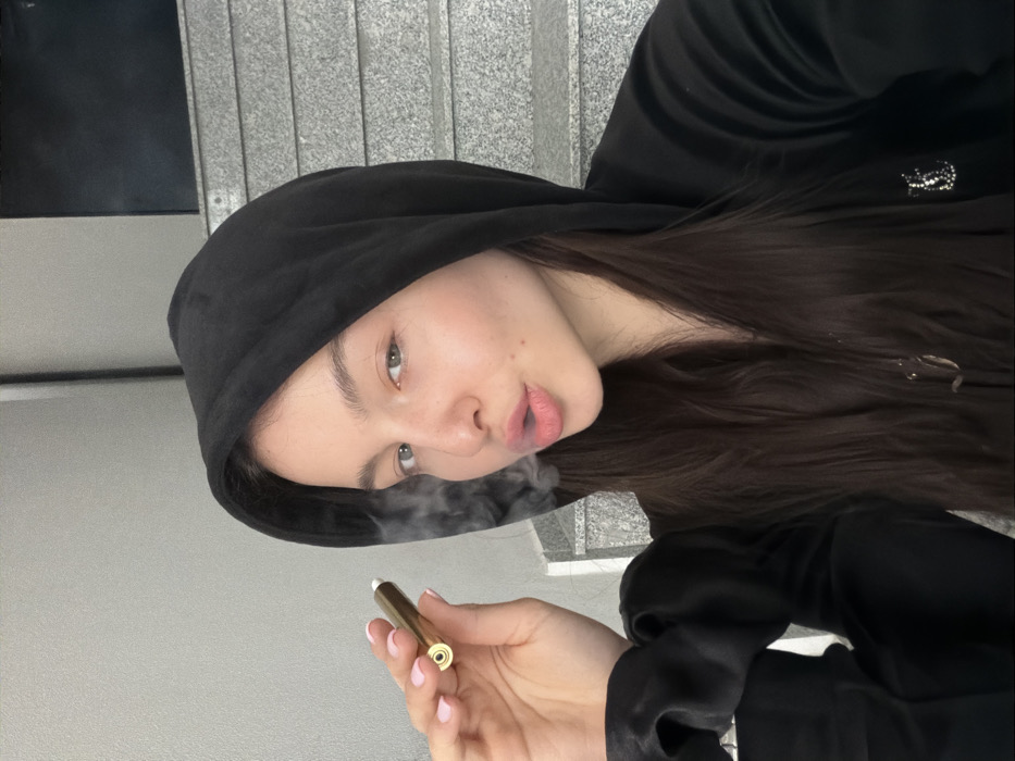 A young adult female, with long dark hair and light-colored eyes, is depicted in a close-up, chest-up selfie. She wears a black hooded sweatshirt with the hood pulled over her head. Her lips are slightly pursed, and she is exhaling a visible plume of white vapor or smoke. In her right hand, adorned with light pink nail polish, she holds a small, cylindrical, metallic object, brass-colored, with a white tip, from which the vapor originates; this object is consistent with a vape pen or e-cigarette designed to resemble a bullet. The background consists of a grey, textured surface with horizontal lines, indicative of concrete or stone steps or panels, suggesting an outdoor or semi-outdoor stairwell. The specific location within Sofia, Bulgaria, is not discernible from the visual evidence provided.