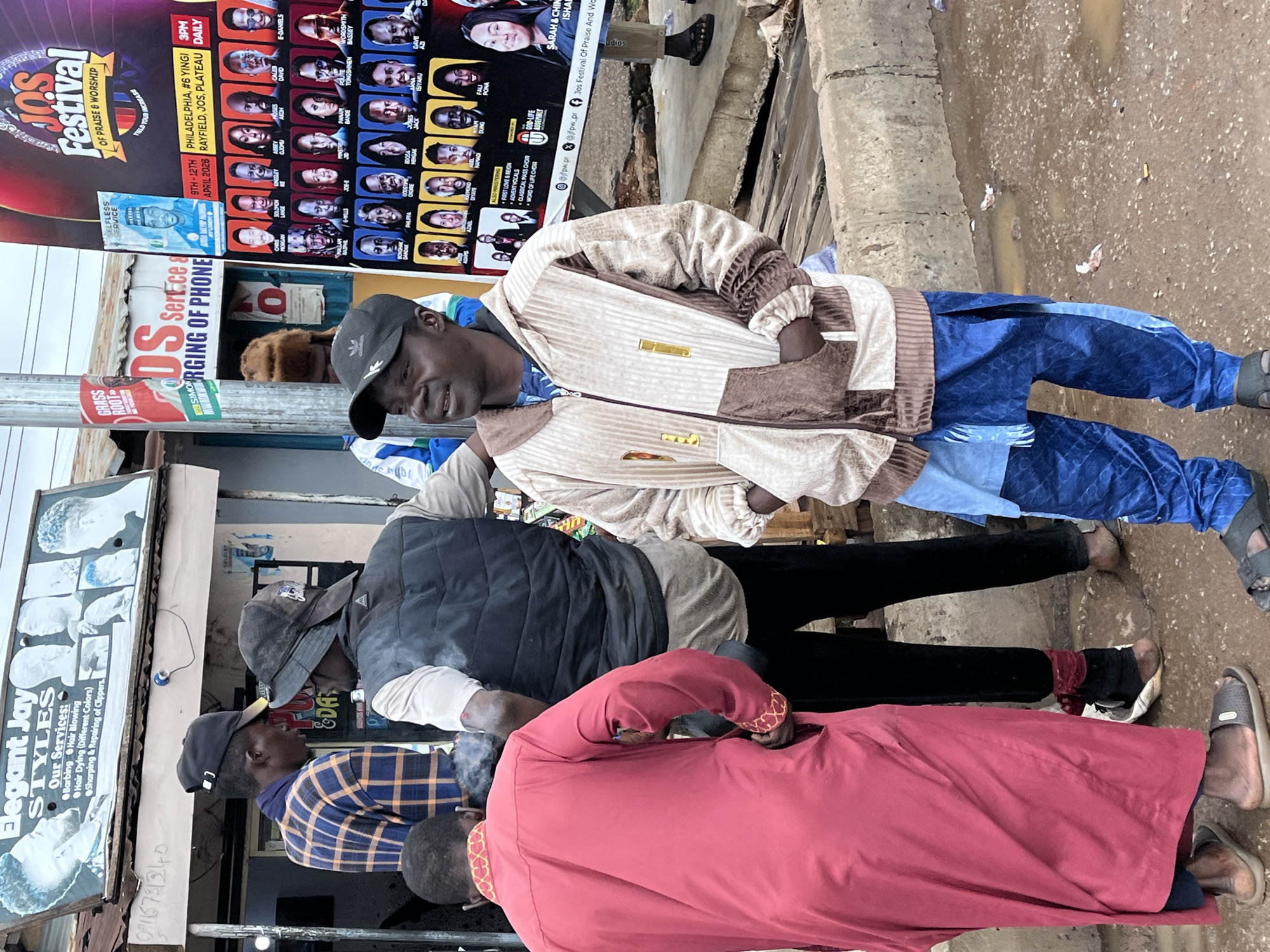 The image captures a street scene in Gura Top, Nigeria, with several individuals standing near a shop and a large poster. In the foreground, a man wearing a beige and brown patterned zip-up jacket and blue patterned pants stands facing left with his hands in his pockets. To his left, a person in a red robe with gold trim stands with their back to the camera. Behind them, two other men are visible; one in a dark vest over a light shirt and black pants, and another in a blue and black plaid shirt and a black baseball cap. A shop sign above them advertises "Elegant Jay Styles" and lists services such as "Barbing," "Hair Blowing," and "Hair Dying." To the right of the men, a large colorful poster for the "JOS Festival of Praise & Worship" is prominently displayed, featuring many portraits of individuals. The ground is wet and muddy, with puddles of water visible, suggesting recent rain. The environment appears to be an outdoor marketplace or street in a Nigerian city.