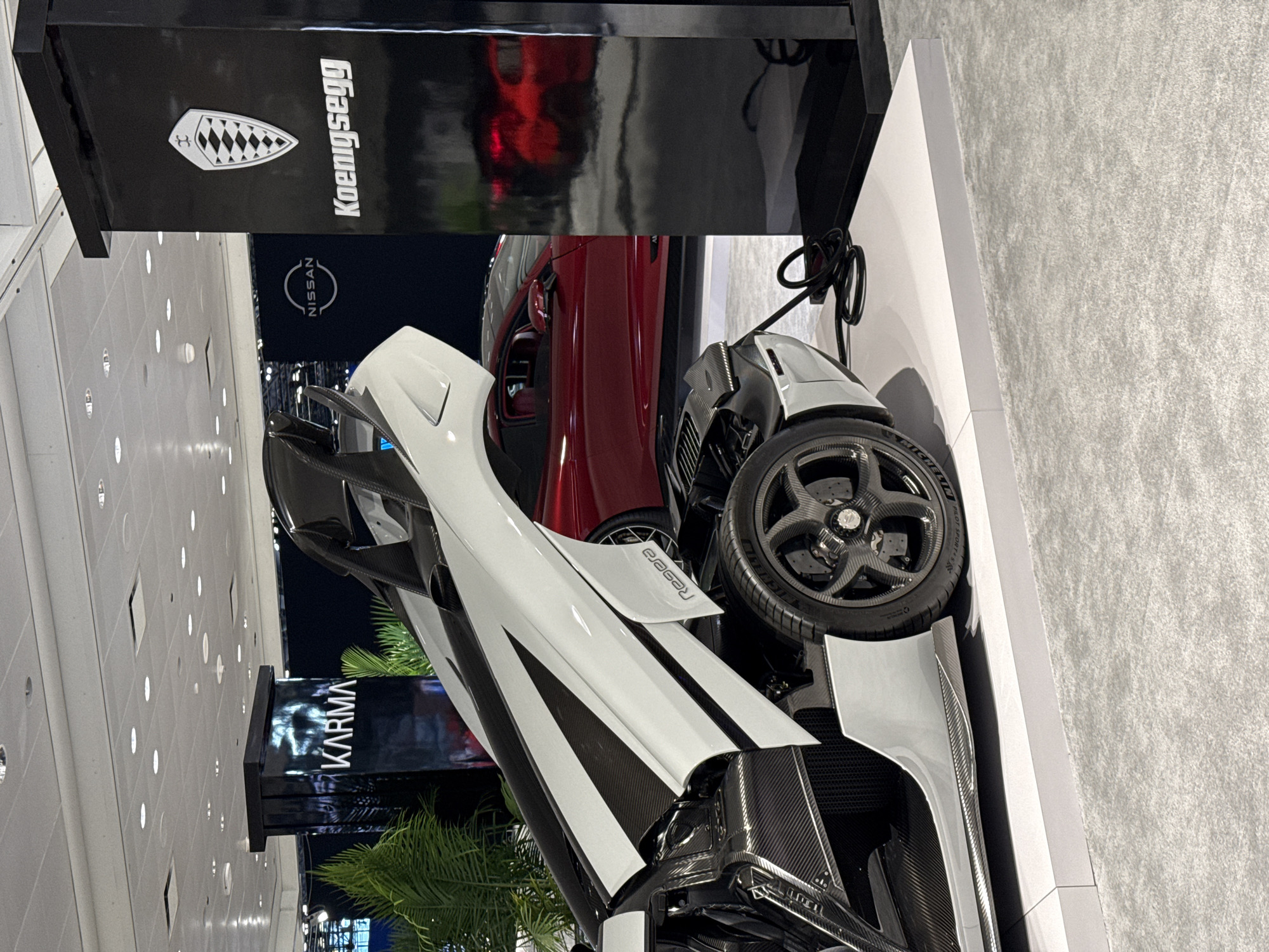 The image captures a car display at an auto show. A Koenigsegg sports car, predominantly white and carbon fiber, is positioned next to a display with the Koenigsegg logo. A red car is partially visible behind the Koenigsegg, and a tire is in clear view. In the background, signs for "NISSAN" and "KARMA" can be seen. The setting is likely an indoor automotive exhibition. No people are directly visible in the frame. It is unclear if the image was taken in New York City.