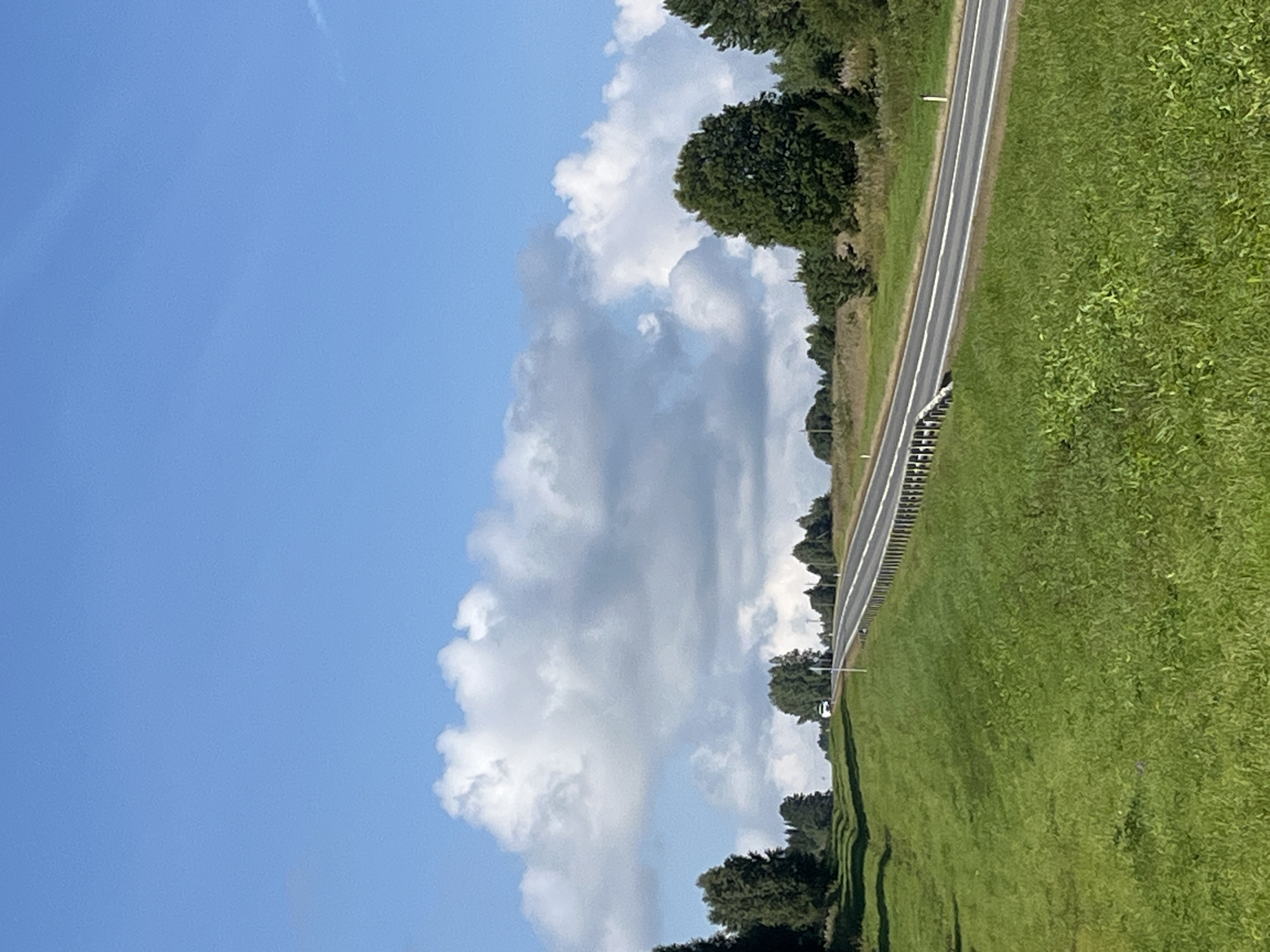 The image captures a rural road in Polozovo, Russia, on a bright, partly cloudy day. A paved road with white lines curves to the right in the mid-ground, bordered by a metal guardrail on the right and a grassy embankment on the left. A white car is visible driving on the road in the distance. The surrounding landscape is characterized by lush green fields and scattered trees. In the background, a line of trees marks the horizon under a blue sky with fluffy white clouds.