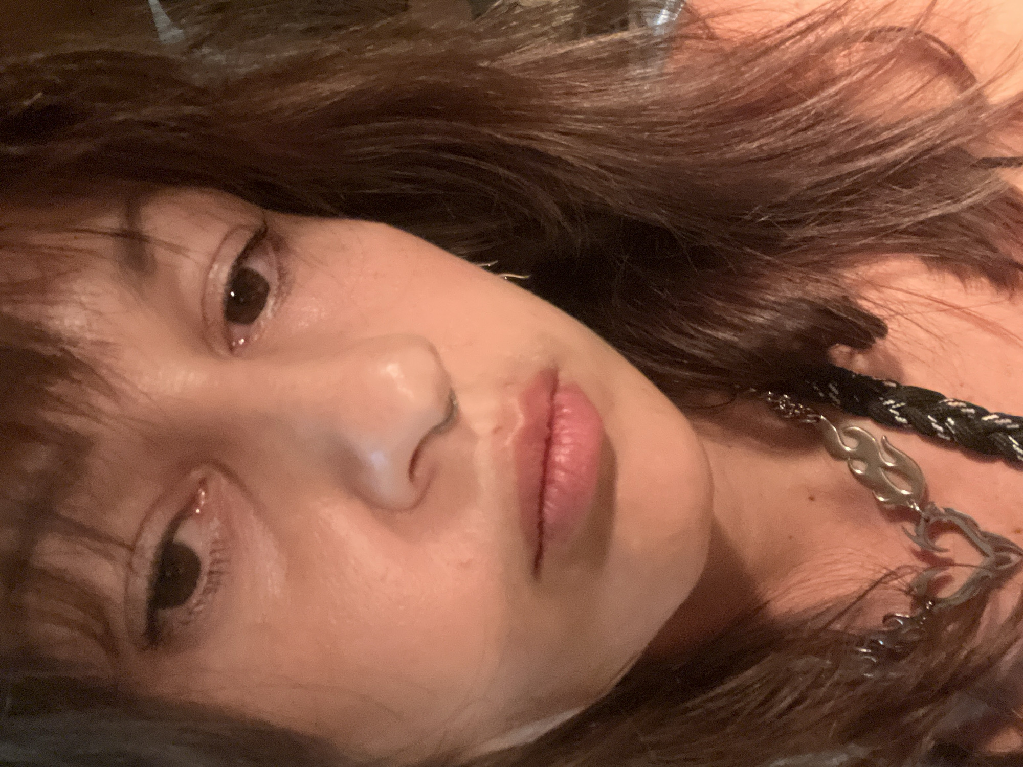 This close-up portrait features a person, likely a young adult female, visible from the chest up, positioned at an angle. She looks directly at the camera with dark brown eyes, her expression calm and neutral. Her lips are natural pink, slightly parted. A subtle nose piercing is visible on her left nostril. Her skin appears smooth with a soft, natural sheen. Dark brown, wavy hair frames her face and cascades around her, suggesting a relaxed posture.
She wears a distinctive necklace featuring several silver-toned, flame-shaped metallic links, which are interspersed with a thick, braided black and white cord. The lighting is soft and warm, suggesting an indoor or low-light environment. The overall mood is serene and introspective. No specific activity beyond posing for the photograph is depicted. There is no visible text in the image. The geographical context for this scene is North Kuta, Indonesia.