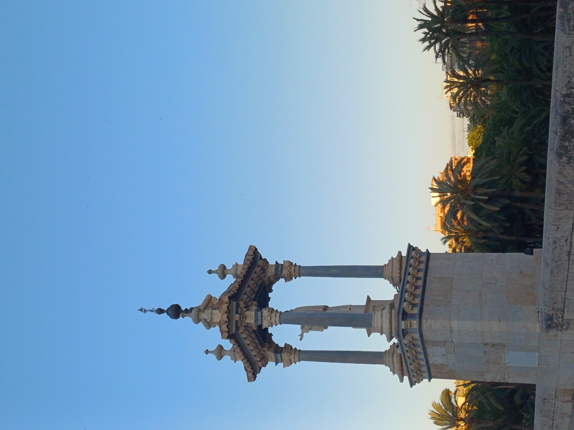 The image depicts a monument or architectural structure set against a clear blue sky in València, Spain. The structure is ornate and made of stone or a similar material, featuring columns and decorative elements. A statue is discernible. Palm trees and buildings can be seen in the background.