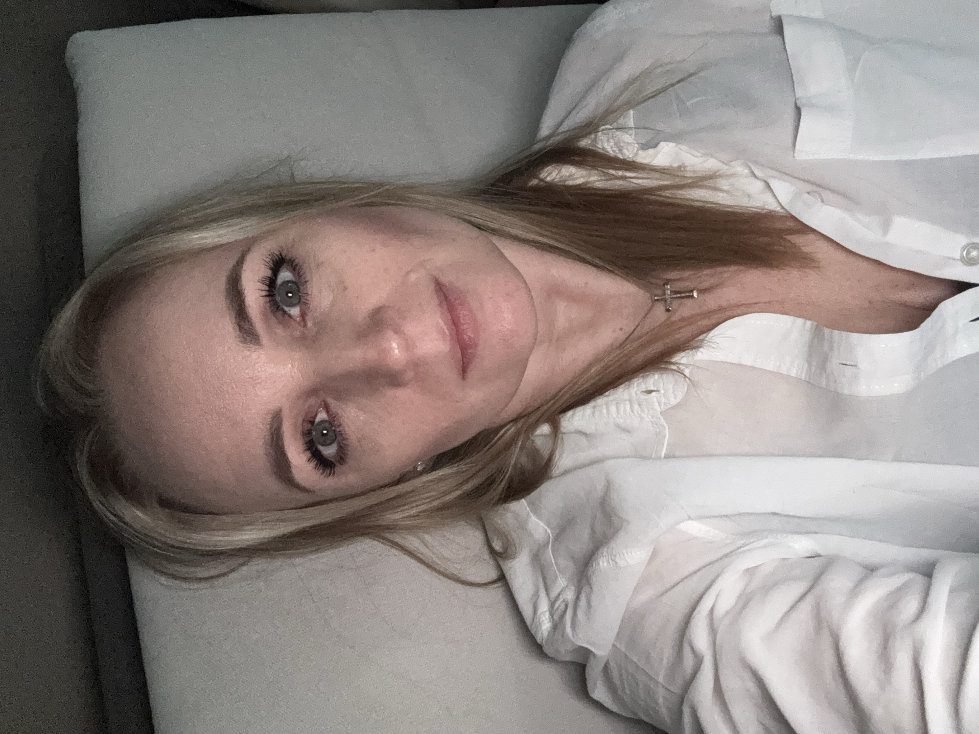 A woman with light skin and light blond hair is lying on a light-colored couch, wearing a white button-down shirt and a silver cross necklace. Her head is turned toward the viewer, and she is looking at the camera. The scene is likely indoors, and there are no details to indicate that it is in Votkinsk, Russia.