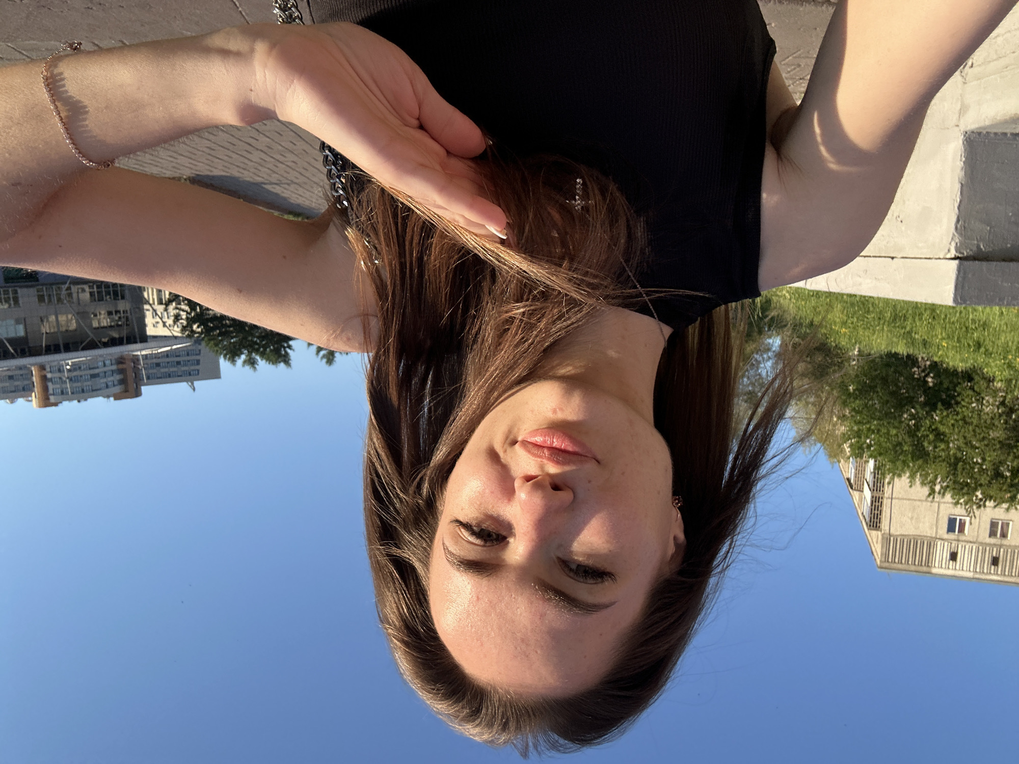 The image features a young woman captured in an outdoor setting, likely a selfie or portrait taken from a low angle. The photograph appears to be inverted, with the subject's head positioned at the bottom of the frame and the sky at the top.
When correctly oriented, the woman looks directly at the camera with a neutral expression, verging on a slight smile. Her eyes appear somewhat squinted, possibly due to bright sunlight. She has long, dark brown hair, parted on the side and flowing around her face and shoulders. Her skin is light, with visible eyelashes (possibly enhanced), dark eyebrows, and natural or light pink lips. Minor blemishes are noticeable on her forehead and chin.
She is wearing a black ribbed tank top or sleeveless shirt. On her left wrist (viewer's right), a delicate, thin bracelet, possibly rose gold, is visible. Her right hand (viewer's left) is raised, with light-colored painted nails, gently touching her hair near her temple. A silver-colored chain is also visible near her right shoulder, possibly part of a necklace or clothing detail.
The background is dominated by a clear, bright blue sky, indicating a sunny day. In the upper corners of the image (when correctly oriented), multi-story buildings and green foliage (trees and grass) are discernible, suggesting an urban or suburban environment. The strong sun creates highlights on her skin and hair. No visible text is present in the media.