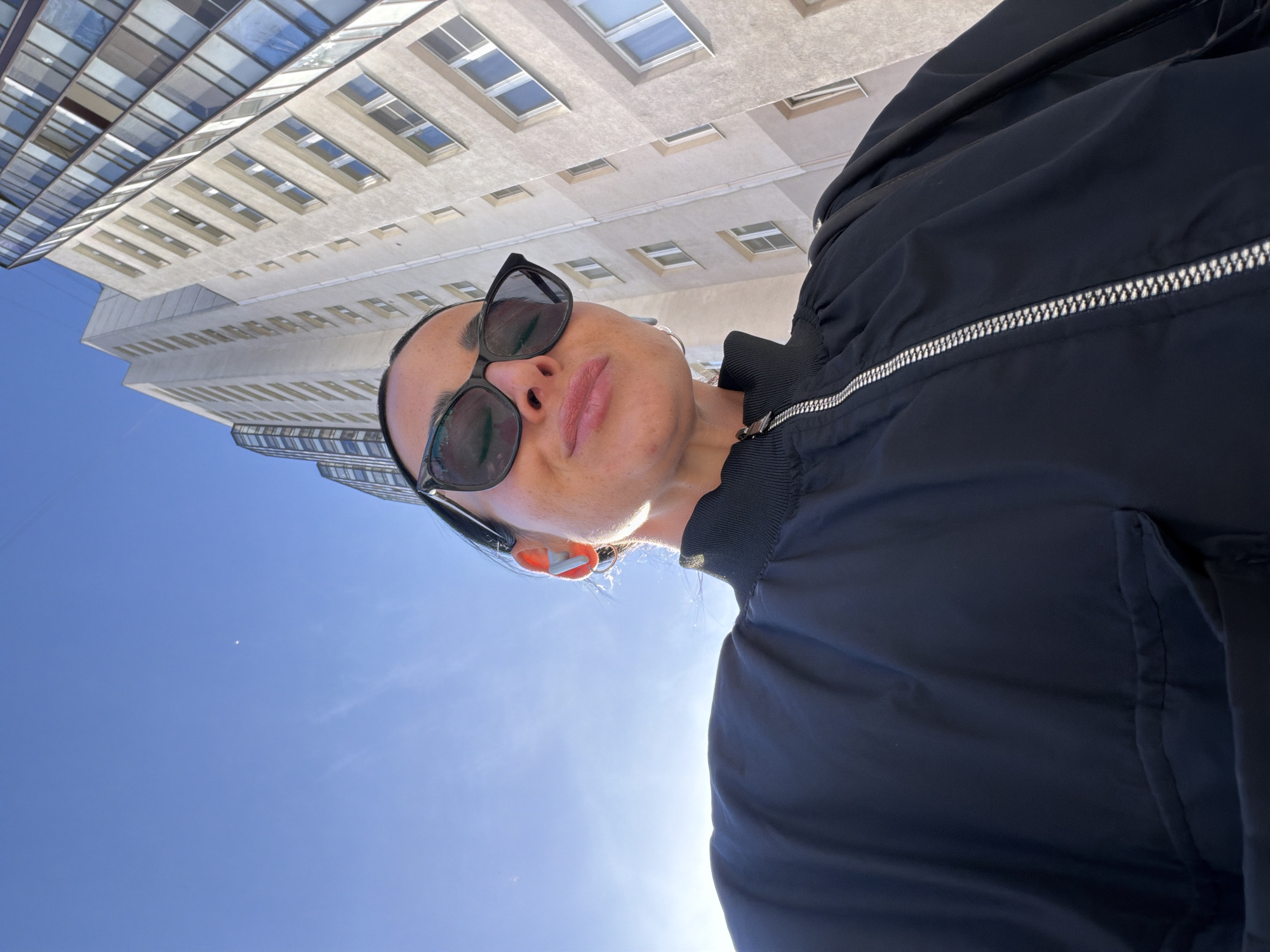 A low-angle shot captures a woman wearing sunglasses and a dark jacket against the backdrop of a blue sky and city buildings. The buildings exhibit a mix of architectural styles with repeating windows, suggesting an urban setting. The sky is clear, indicating a sunny day. The woman's face is well-lit, and she appears to be looking up at the camera. The overall composition gives a dynamic perspective of a person in an urban environment. Without more information, I am unable to verify the location.