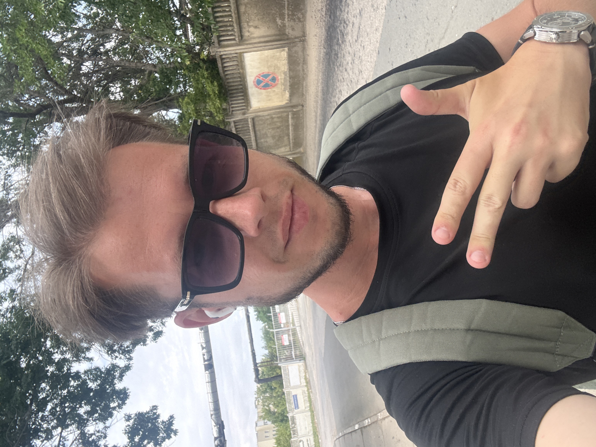 A man with light brown hair, sunglasses, and a short beard is taking a selfie outdoors. He is wearing a black t-shirt and a light olive-green backpack. His right hand is raised, with his index, middle, and ring fingers extended, and his thumb tucked in. He is wearing a silver watch on his left wrist. In the background, there is a concrete wall with a circular sign depicting a red circle with a blue cross through it, indicating a no-parking zone. Trees and buildings are visible further in the background, along with what appears to be industrial piping overhead. The location is identified as Aeroflotsky, Ukraine.