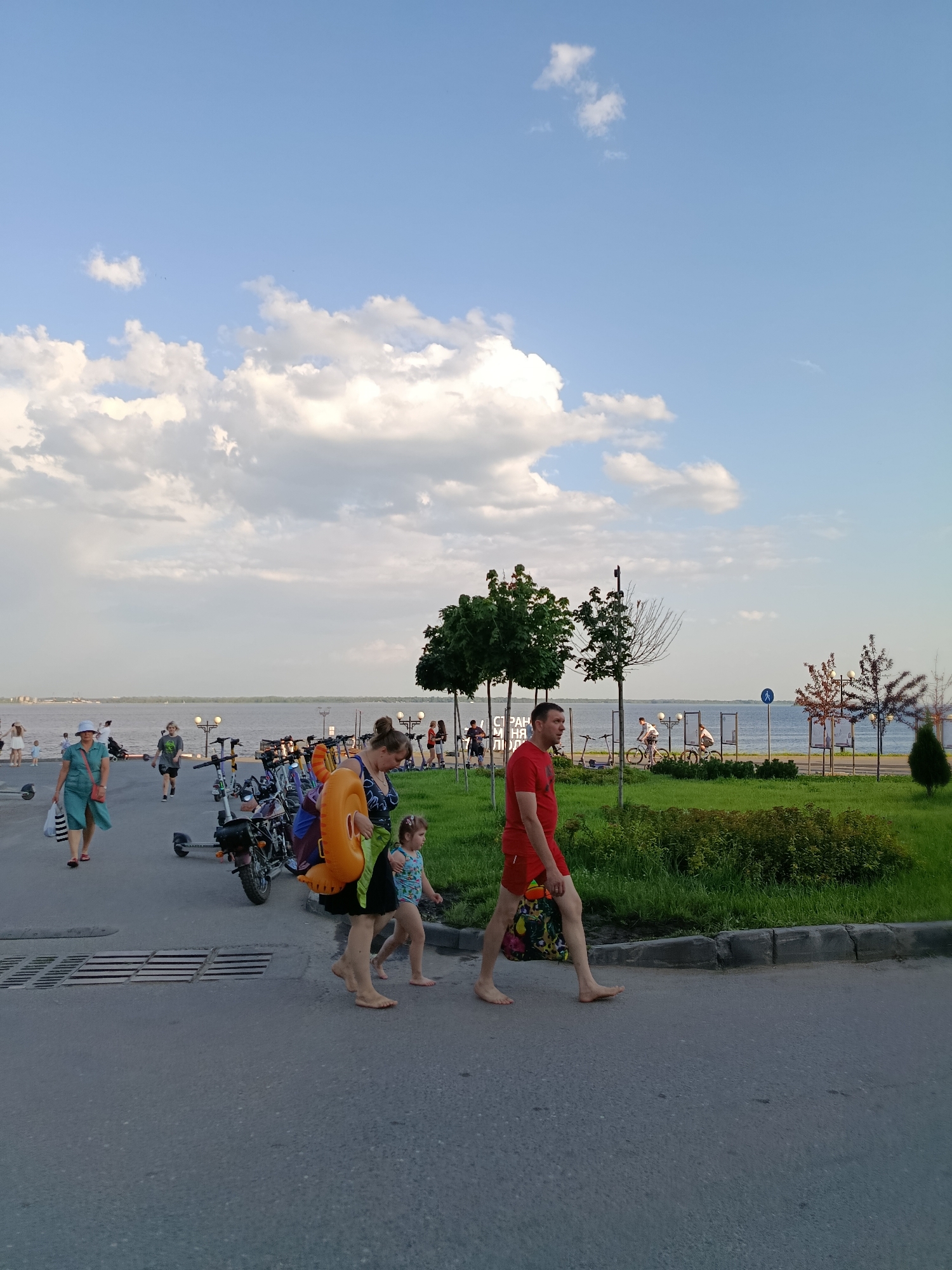 The image captures a scene along the Volga River embankment in Saratov, Russia. A group of people are walking on the asphalt pathway. In the foreground, a man in a red t-shirt and red shorts walks barefoot, carrying a colorful bag. To his left, a woman in a black and blue patterned top and a black skirt walks barefoot, holding a bright orange inflatable ring and a green bag. A young girl in a blue and white swimsuit walks beside her. Further to the left, a woman in a teal dress and a straw hat walks with shopping bags. In the background, several bicycles and electric scooters are parked along the embankment. The Volga River stretches across the horizon, with a cloudy sky above. Green grass and trees line the embankment, creating a pleasant recreational area.