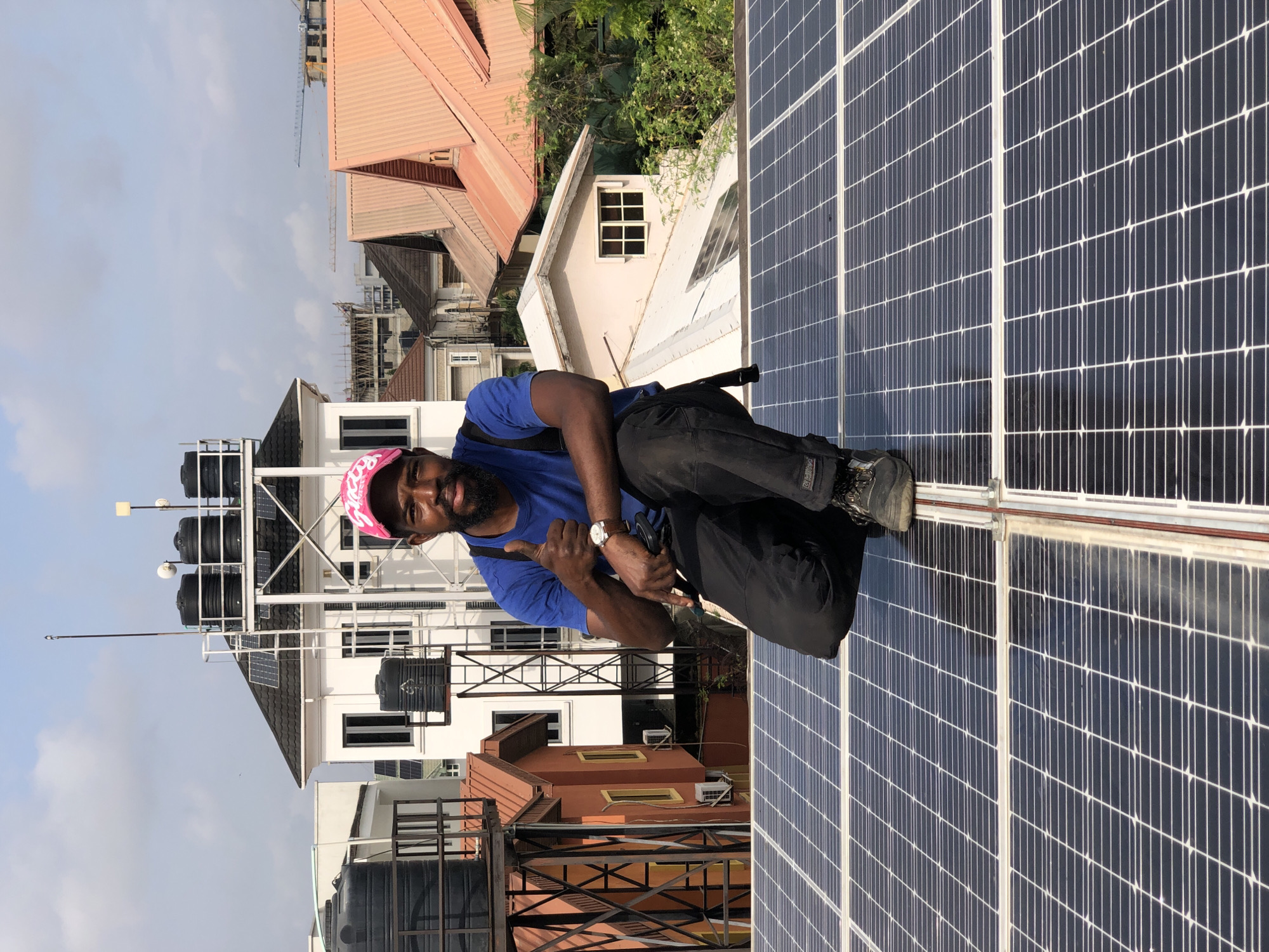 In Ilado, Nigeria, a man in a blue t-shirt, black pants, and a pink cap is kneeling on a rooftop covered with solar panels. He is giving a thumbs-up gesture. The man is in the foreground, with several buildings and a clear sky visible in the background. The buildings have various roof types and some appear to be under construction. Water tanks and antenna structures are also present on some rooftops, indicating a densely populated urban environment.