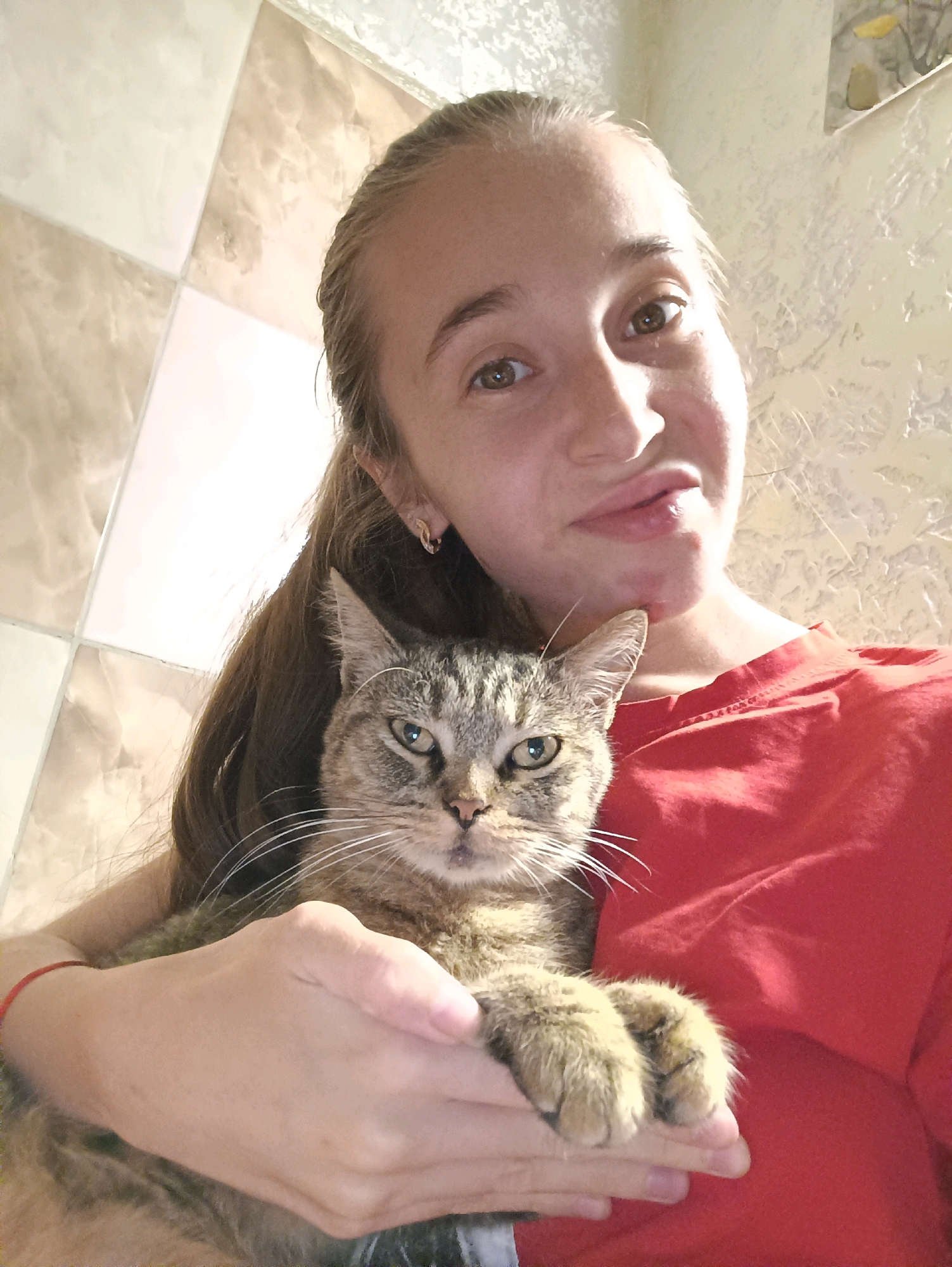 In a self-portrait taken indoors, a young woman with long brown hair braided to her right side is holding a tabby cat. The woman is wearing a red t-shirt and a thin red bracelet on her left wrist. She has brown eyes and is looking towards the camera with her lips slightly parted. The cat, also looking at the camera, is being held in her arms, with its paws resting on her hand. The background features a tiled wall with a diagonal pattern and a textured wall on the right. There is no indication of the city of Vypasne, Ukraine in the image.