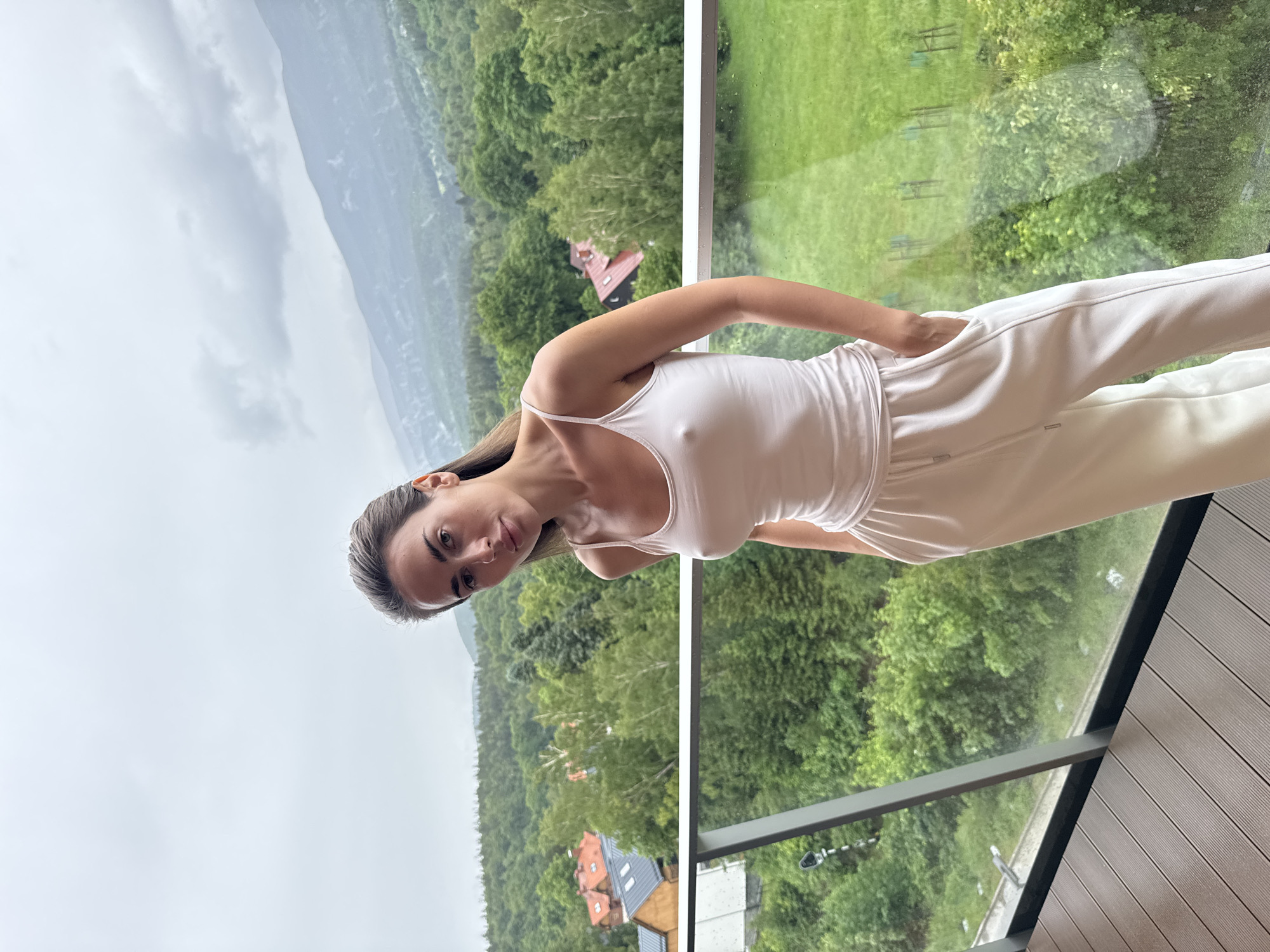 A portrait-oriented image, originally rotated 90 degrees counter-clockwise, depicts a woman standing on a balcony with a panoramic natural landscape behind her. The woman, with long dark hair, is wearing a white spaghetti-strap tank top and matching white sweatpants or joggers. She is looking directly at the camera with a neutral to slightly smiling expression. Her right hand is casually placed in her pocket.
The background showcases a dense green forest covering undulating terrain, suggesting mountains or large hills. Several houses with distinct reddish-brown and grey roofs are nestled among the trees in the distance. The sky above the mountains is overcast with heavy grey clouds, indicating cloudy weather. The balcony features a glass railing, which reflects some of the green landscape and appears to have slight moisture or streaks, possibly from recent rain. The floor of the balcony is made of dark, horizontally striped composite decking. No visible text is present in the image.