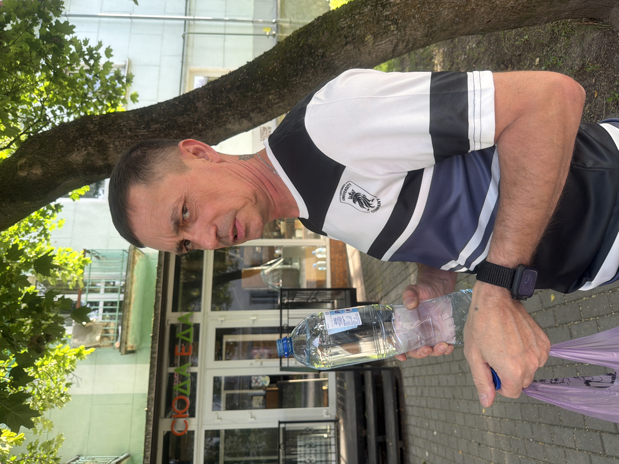 A man stands outdoors in front of a light green building with a storefront. He is holding a clear plastic bottle of water in his right hand and a purple plastic bag in his left. He wears a black and white striped t-shirt with a crest on the left side of his chest and a black watch on his left wrist. The building appears to be a commercial establishment with large glass doors and windows. The storefront signage is partially obscured but appears to read "СЮДА ЕДА" in orange and green letters. The ground is paved with bricks. A tree with a rough bark trunk is to the right of the man, with green leaves overhead. The lighting suggests it is daytime, possibly sunny, with dappled light filtering through the leaves. The background shows more of the building, including a balcony with a green railing, and possibly other buildings or structures. The overall setting is an urban environment.