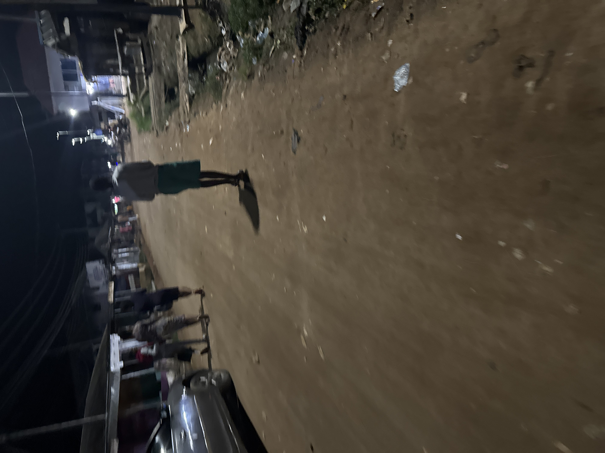 The image depicts a street scene in Ihiagwa, Nigeria. A person wearing a light-colored shirt and green shorts is walking along the side of a dirt road. The road surface is uneven and littered with debris. Buildings are visible along the side of the road, with some appearing to be shops or stalls. A vehicle is parked along the roadside, partially obscuring the view. Other people and objects are also visible in the background. The lighting suggests it is either late evening or early morning.