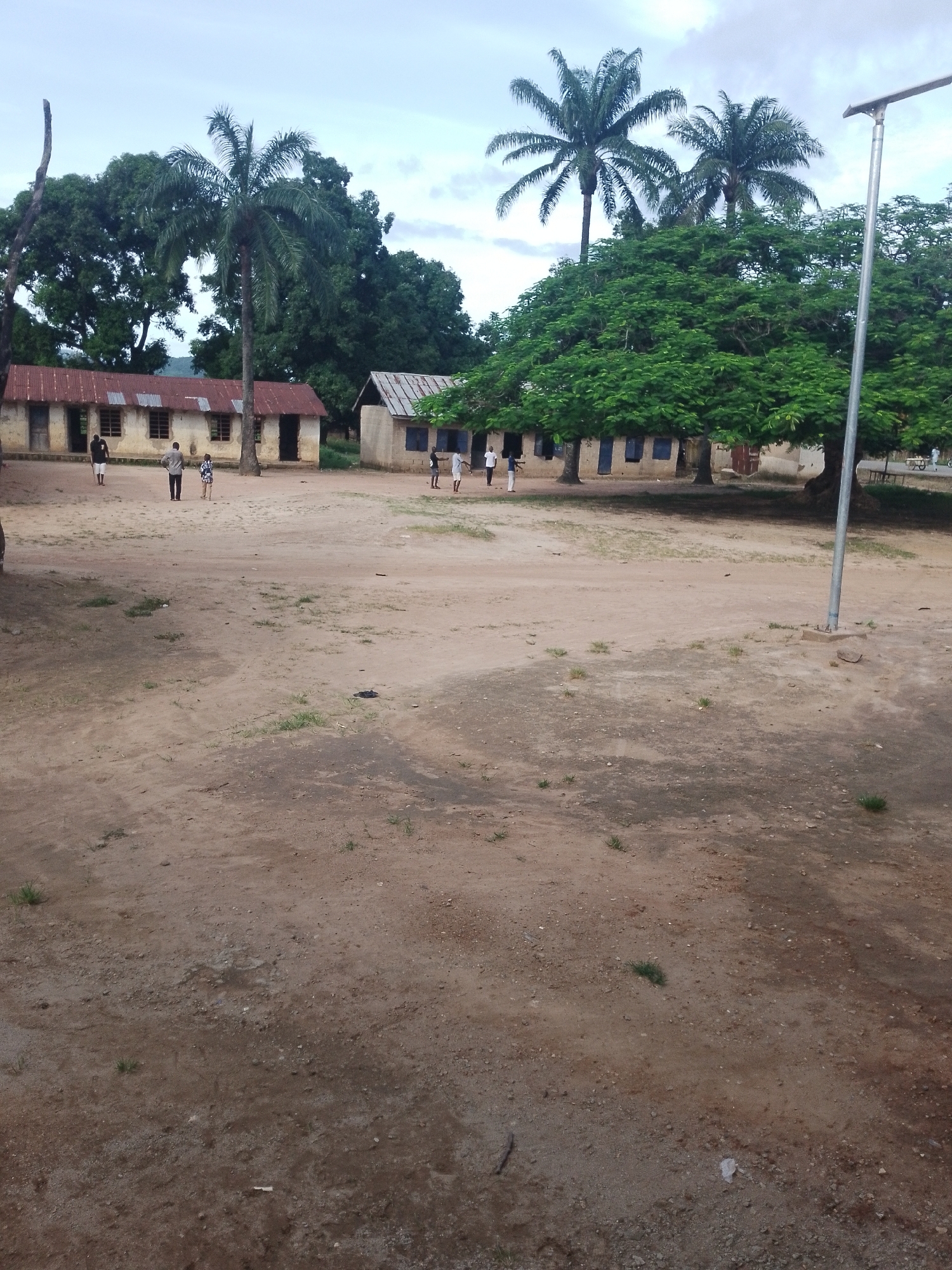 An outdoor, medium-shot, eye-level photograph captures a dusty, open area with buildings and trees. The ground is composed of dry, reddish-brown dirt with sparse patches of green grass and small stones. In the background, two single-story buildings with corrugated metal roofs and light-colored walls stand side-by-side. The building on the left has several open doorways and windows, while the building on the right has blue-shuttered windows. Several people, appearing to be children or young adults, are scattered around the area in front of the buildings. One person in the foreground stands facing away from the camera, wearing a dark shirt and white shorts. Another person, wearing a light-colored collared shirt and dark pants, stands with their back to the camera. A child in a patterned dress stands near them. Further to the right, more individuals are visible, some standing and some walking. A tall palm tree stands to the left of the central building, and a large tree with dense green foliage dominates the right side of the frame, with two more palm trees visible behind it. A light-colored, modern-looking pole with a solar panel attached to its top stands on the right side of the image. The sky above is overcast with diffused light.